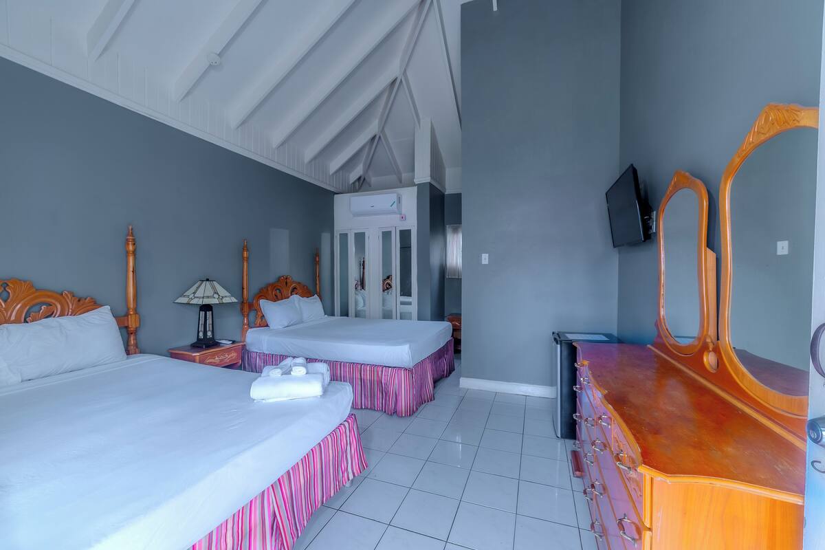 Une chambre d’hôtel avec deux lits, une commode en bois, une télévision et un ventilateur de plafond.