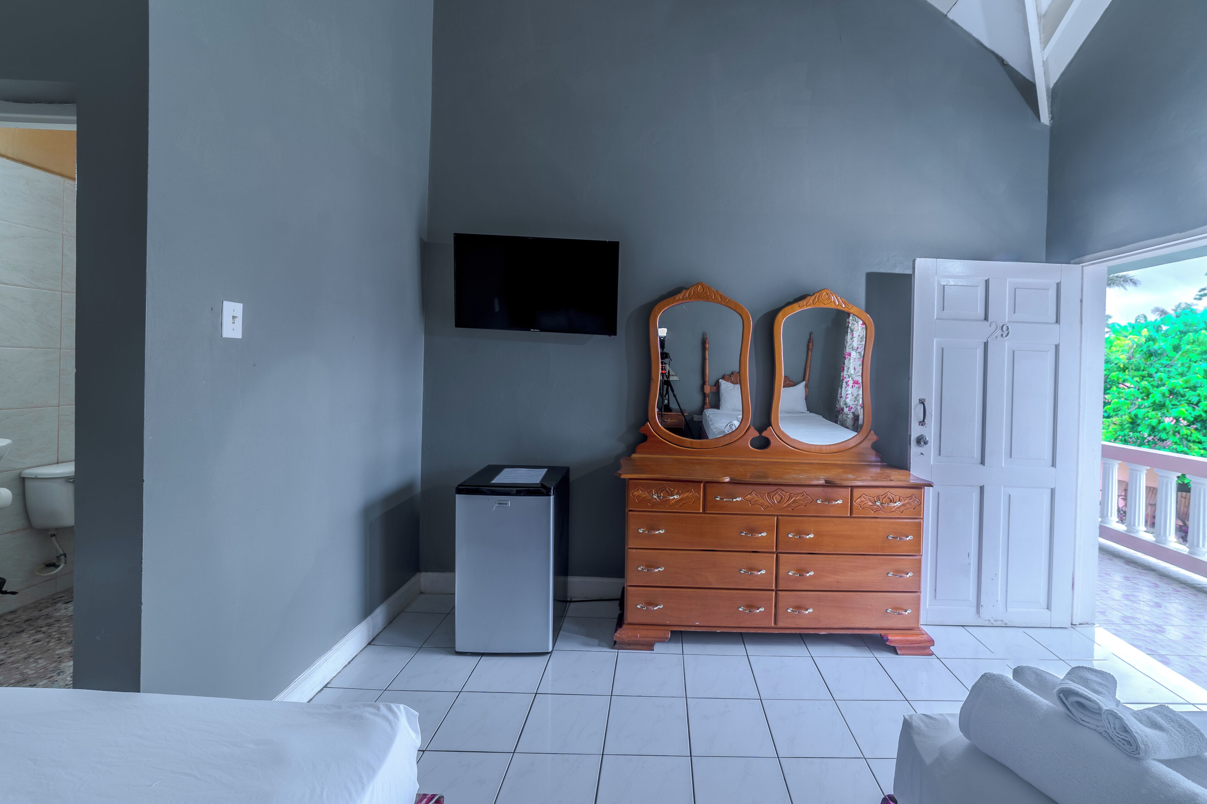 Ein Schlafzimmer mit Bett, Schminktisch, Fernseher, einer Tür und einem Balkon mit Grünpflanzen.
