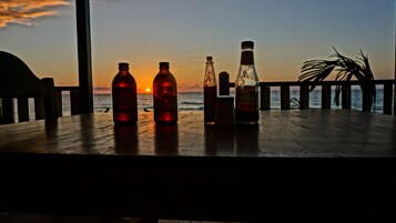 Una mesa de madera con botellas y una copa en un balcón con vista al océano al atardecer.