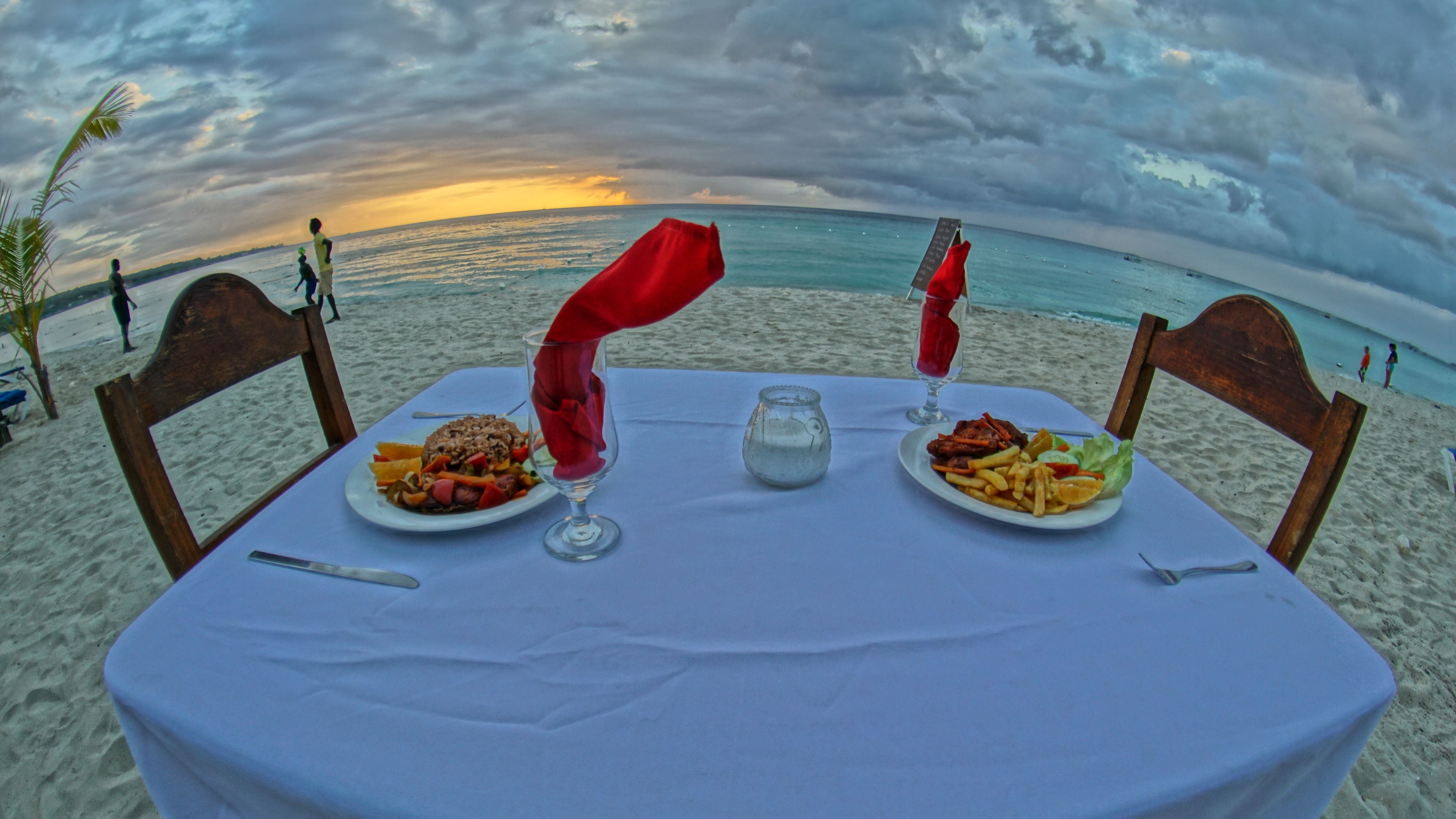 Une table dressée sur une plage, avec deux assiettes de nourriture, un verre d’eau et une bougie.