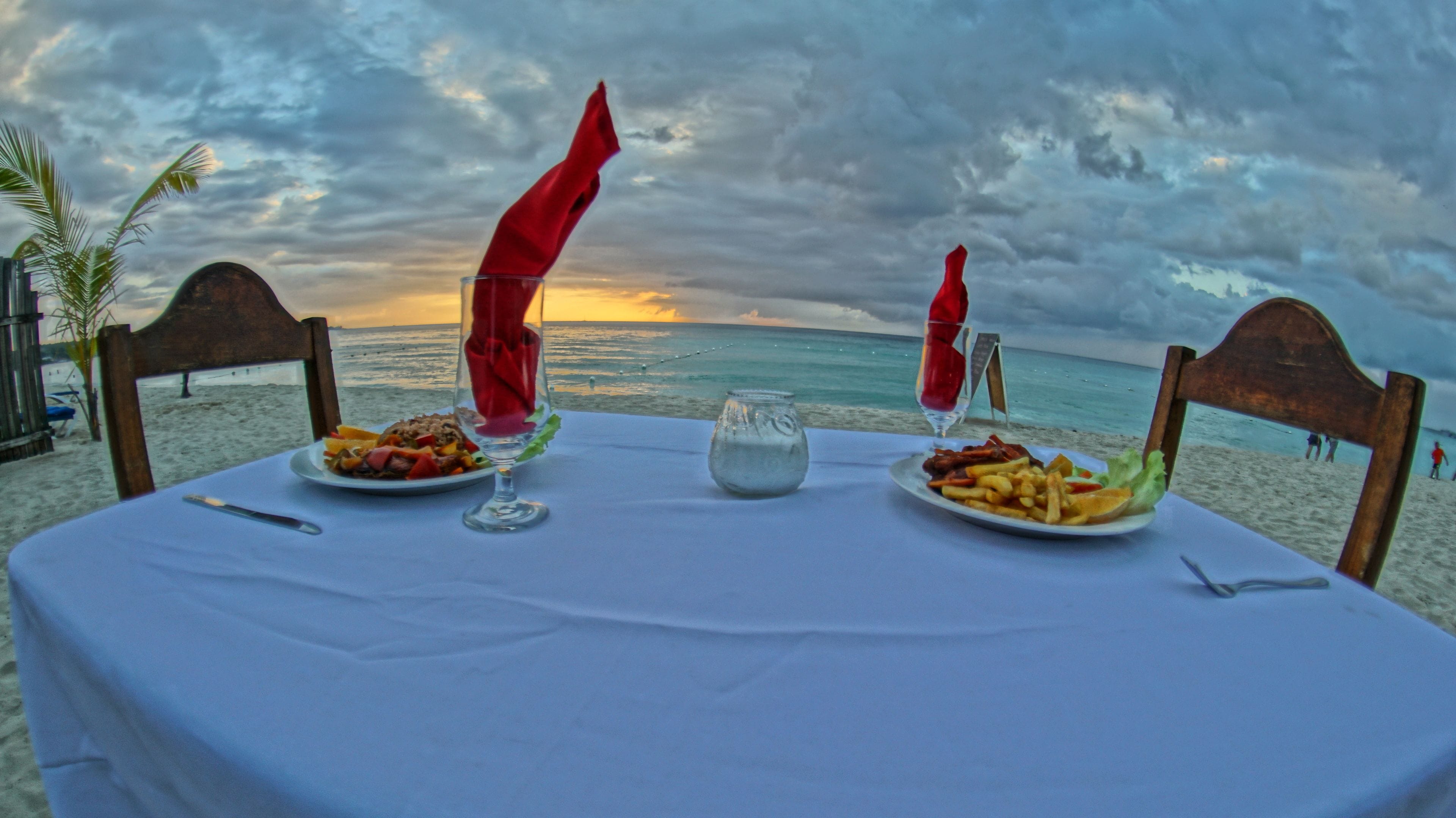 Tavola imbandita per due, con piatti di cibo e bicchieri, con vista su una spiaggia e un cielo nuvoloso.