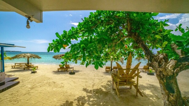 A beachside area with wooden lounge chairs, a thatched umbrella, and a tree providing shade. - Jamaica Tamboo Resort (Negril)