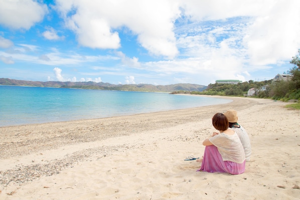 Una mujer sentada en una playa arenosa, con vista clara al mar y colinas distantes.
