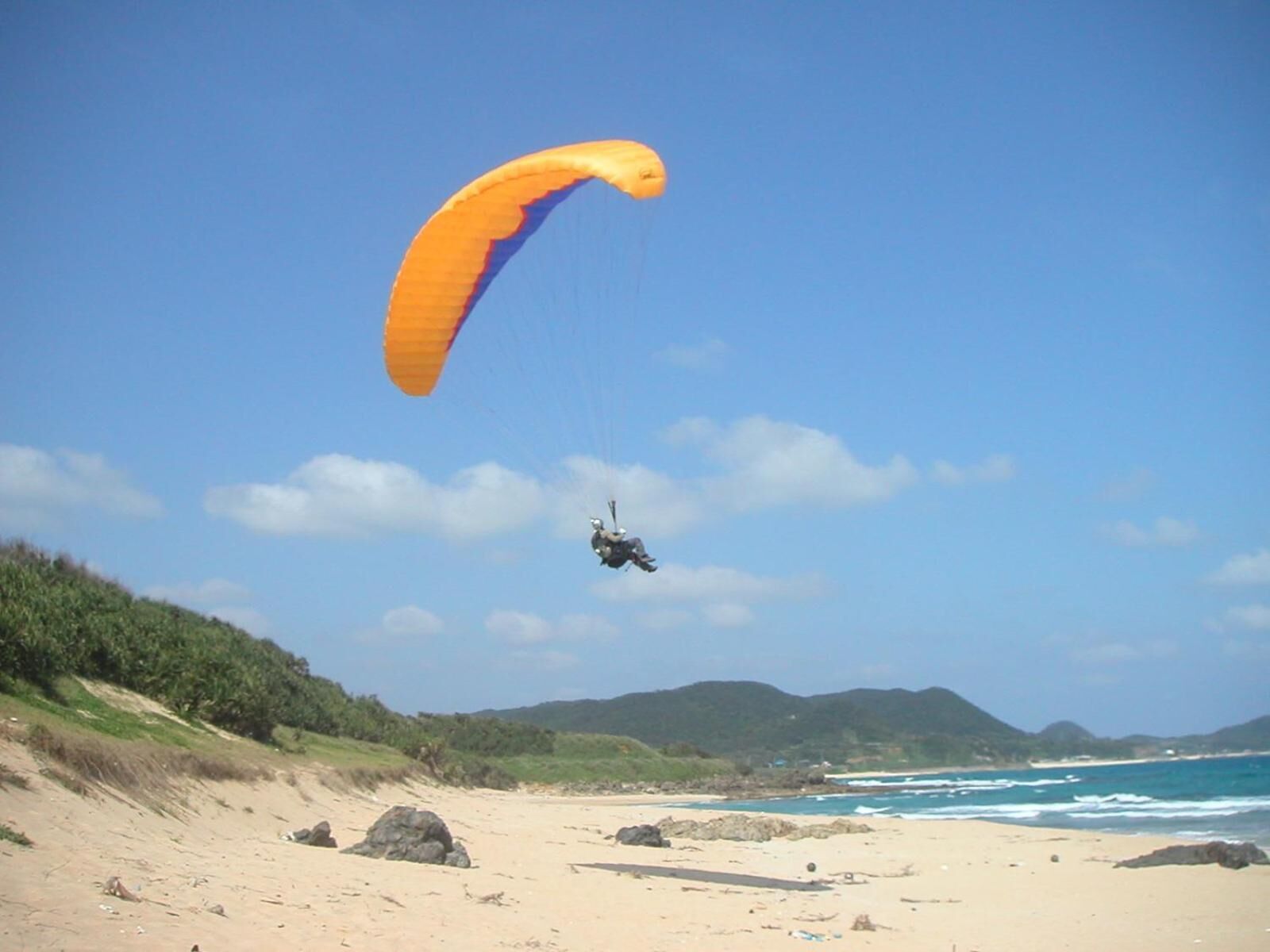 A person paragliding over a beach with a clear sky and distant mountains.
