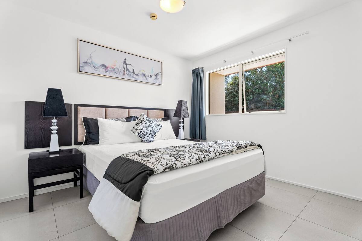 A modern bedroom with a large bed, two bedside tables, a ceiling light, a window with curtains, and a wall-mounted artwork.