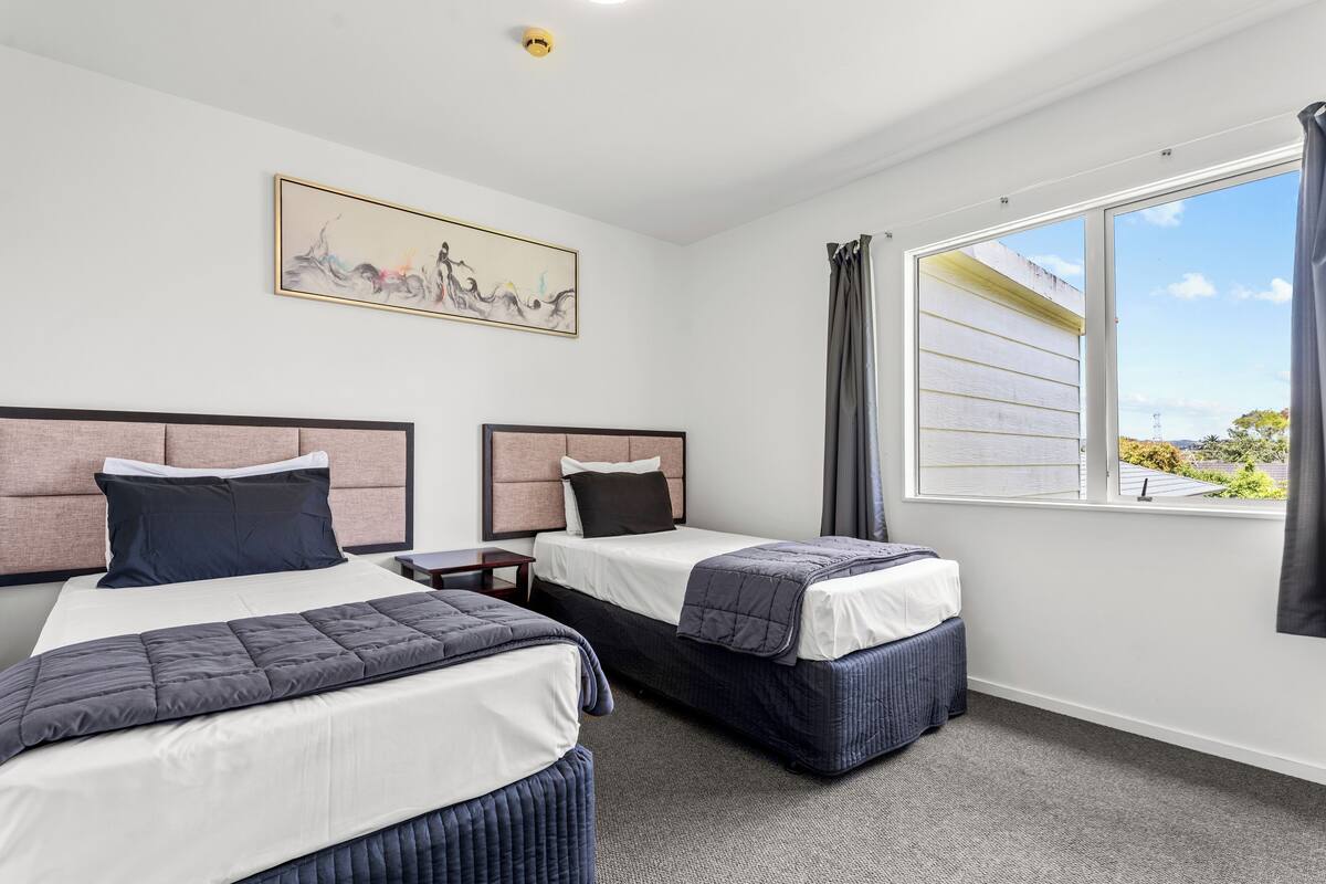 A hotel room with two beds, a large window with curtains, a painting on the wall, and a ceiling light.