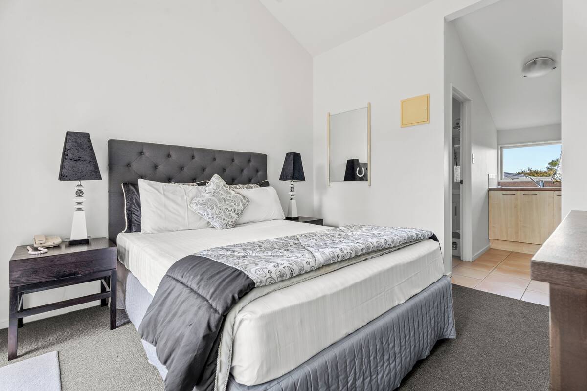 A modern bedroom with a large bed, two bedside tables, a nightstand, a mirror, and a view of the kitchen and balcony.