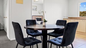 A dining area with a round table and four dark chairs, a potted plant on the table, and a view of a kitchen and a window with a view of trees. - Avondale Motor Inn (Auckland)