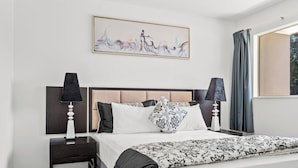 A neatly arranged hotel room with a large bed, two bedside tables, a headboard, a wall-mounted artwork, and a window with curtains. - Avondale Motor Inn (Auckland)