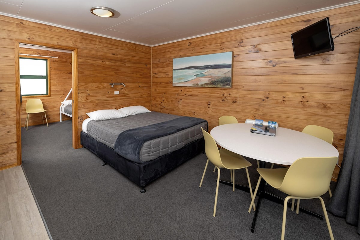 Une pièce comprenant un lit, une table avec des chaises, un téléviseur fixé au mur et un tableau.
