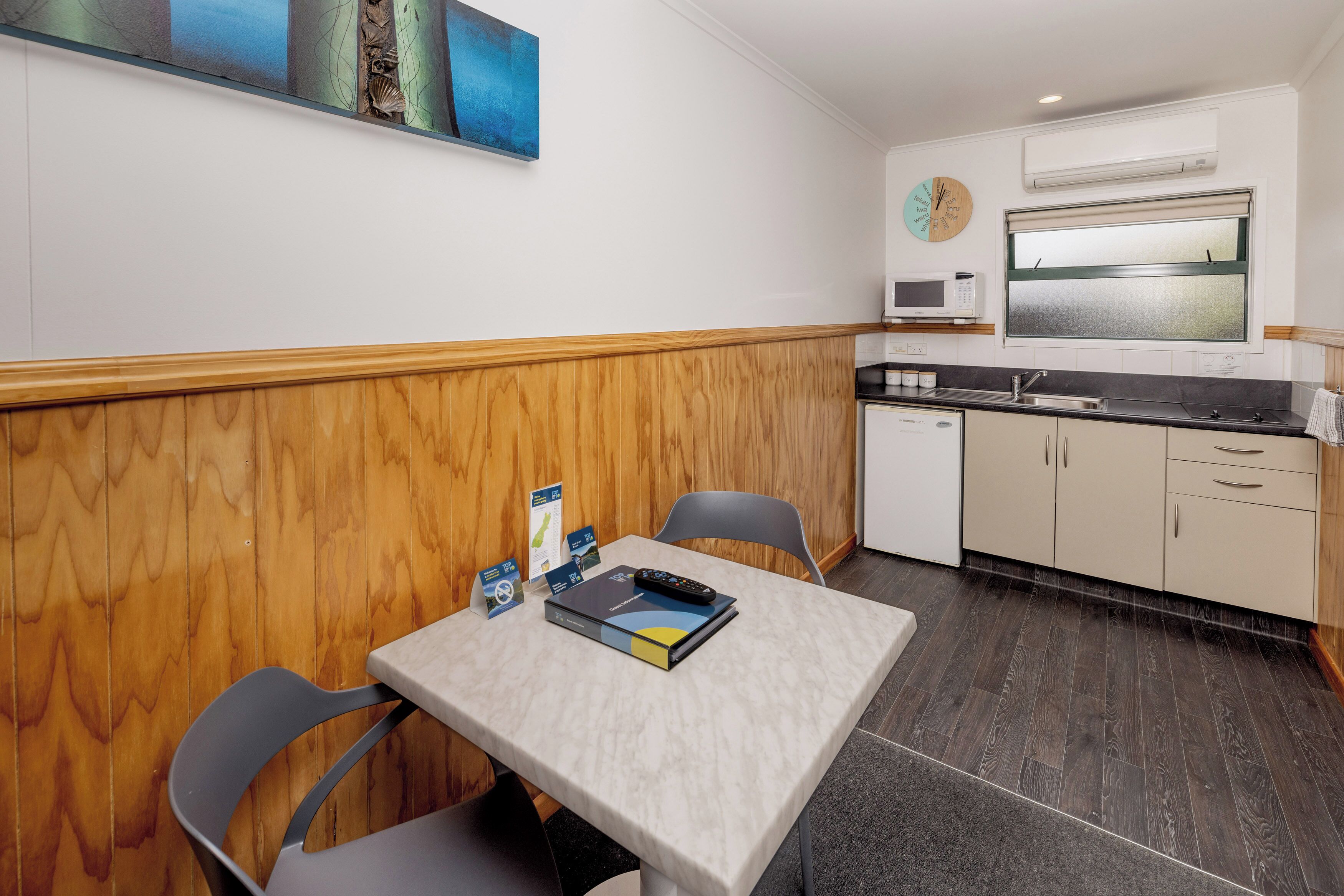 Una cocina compacta con microondas, pileta y alacenas. Hay una mesa de comedor con sillas y un pequeño cuadro en la pared.