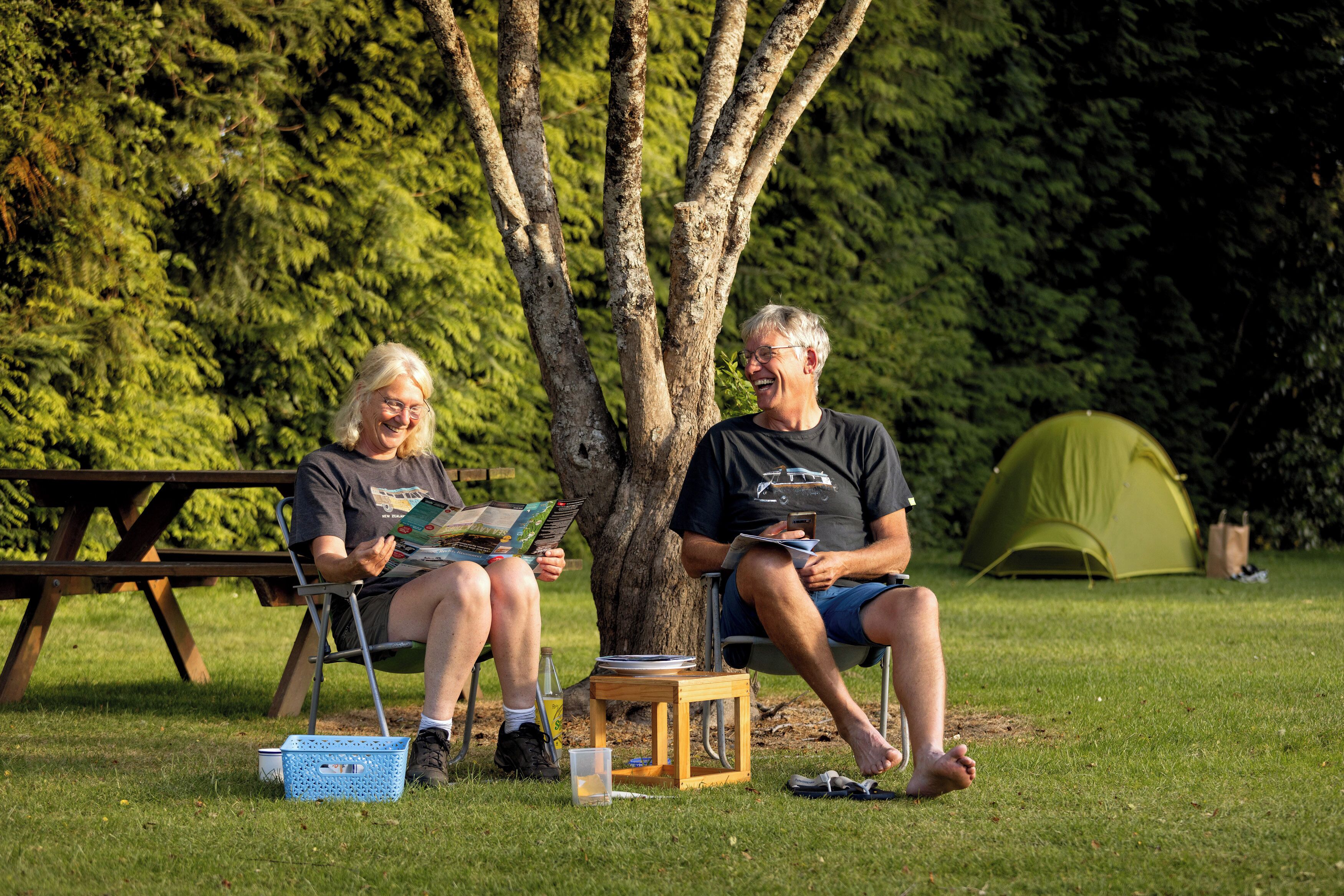 Dos personas sentadas en sillas bajo un árbol, con una mesa de picnic y una tienda de campaña al fondo.