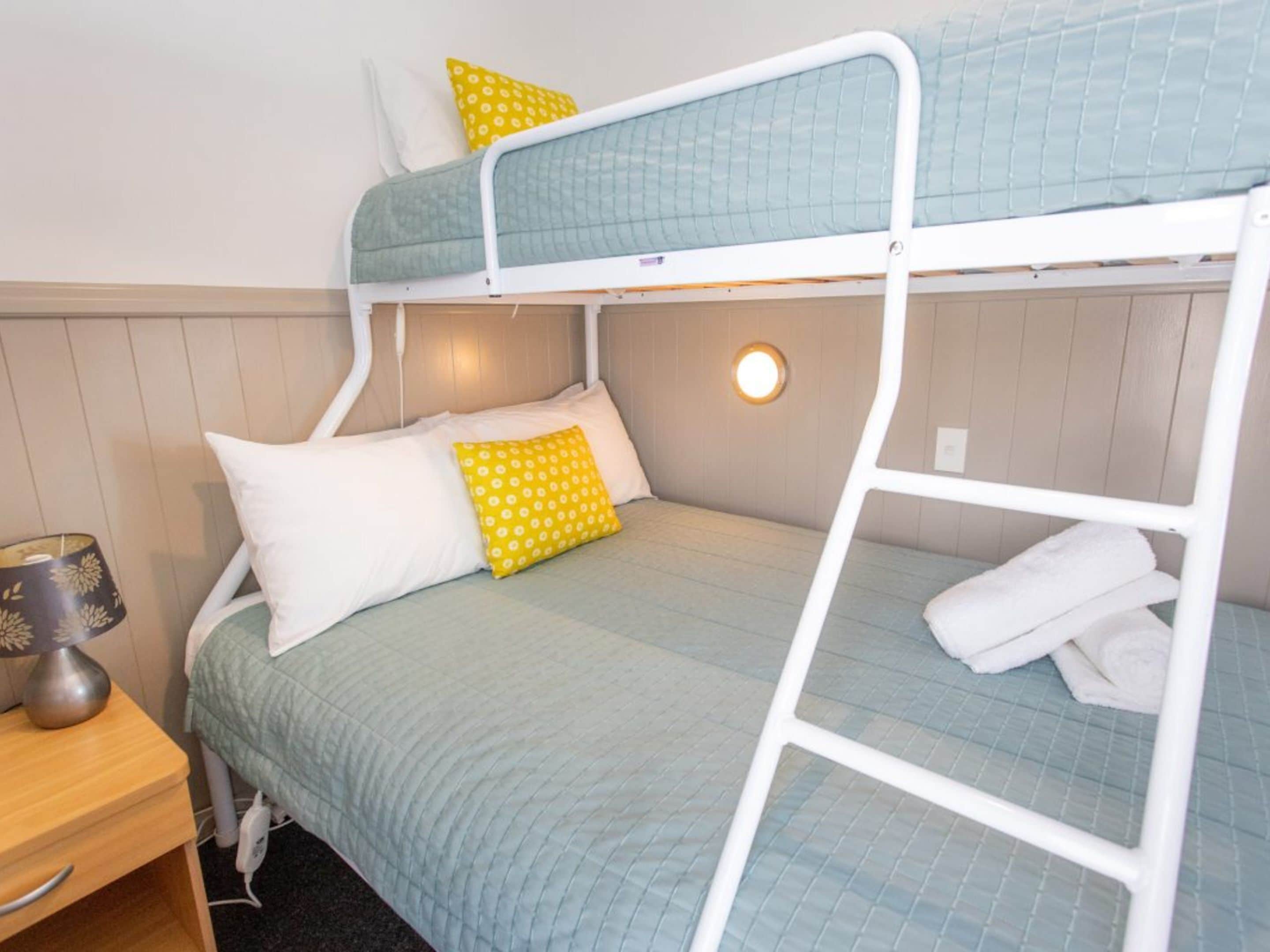 A bunk bed with a green quilt, white pillows, and a yellow pillow with a floral pattern. There is a bedside table with a lamp and a folded towel on the lower bunk.