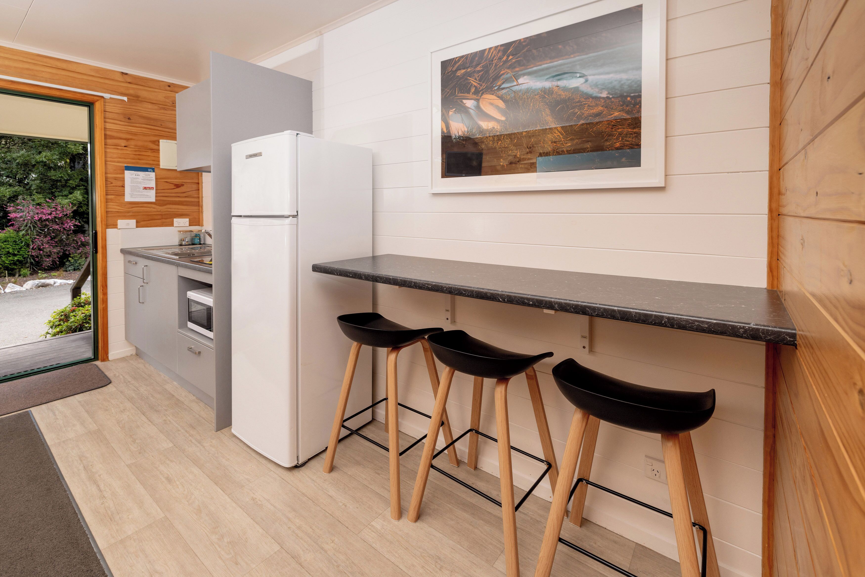 Una cocina moderna con un refrigerador blanco, un microondas y una encimera negra. Hay tres taburetes negros con patas de madera. Encima de la encimera, se encuentra un cuadro enmarcado colgado en la pared.