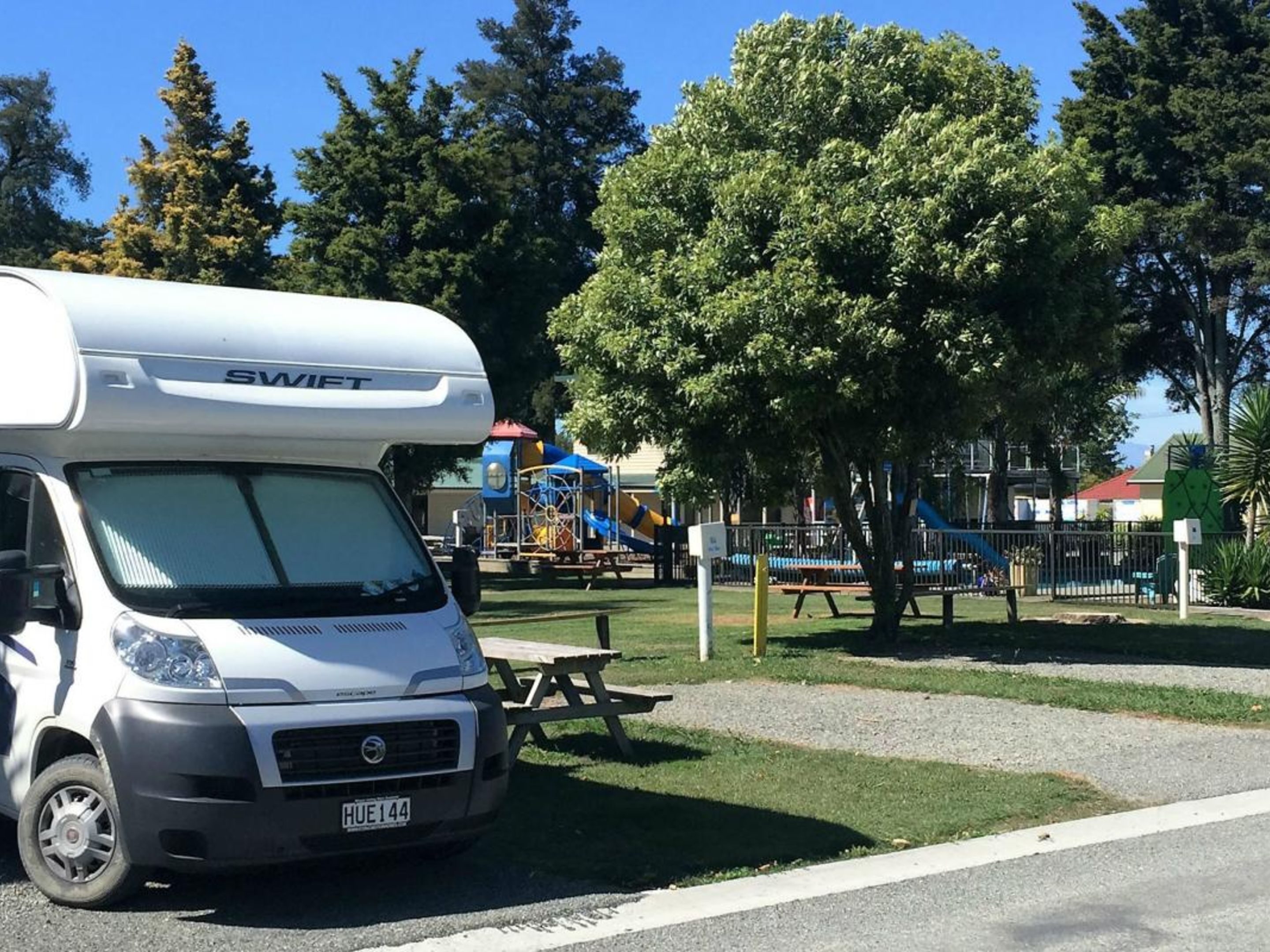 Una caravana blanca con el nombre de marca 'SWIFT' estacionada cerca de un parque infantil con mesas de picnic y una zona vallada.