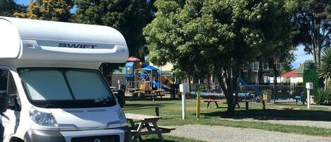 Una caravana blanca con el nombre de marca 'SWIFT' estacionada cerca de un parque infantil con mesas de picnic y una zona vallada.