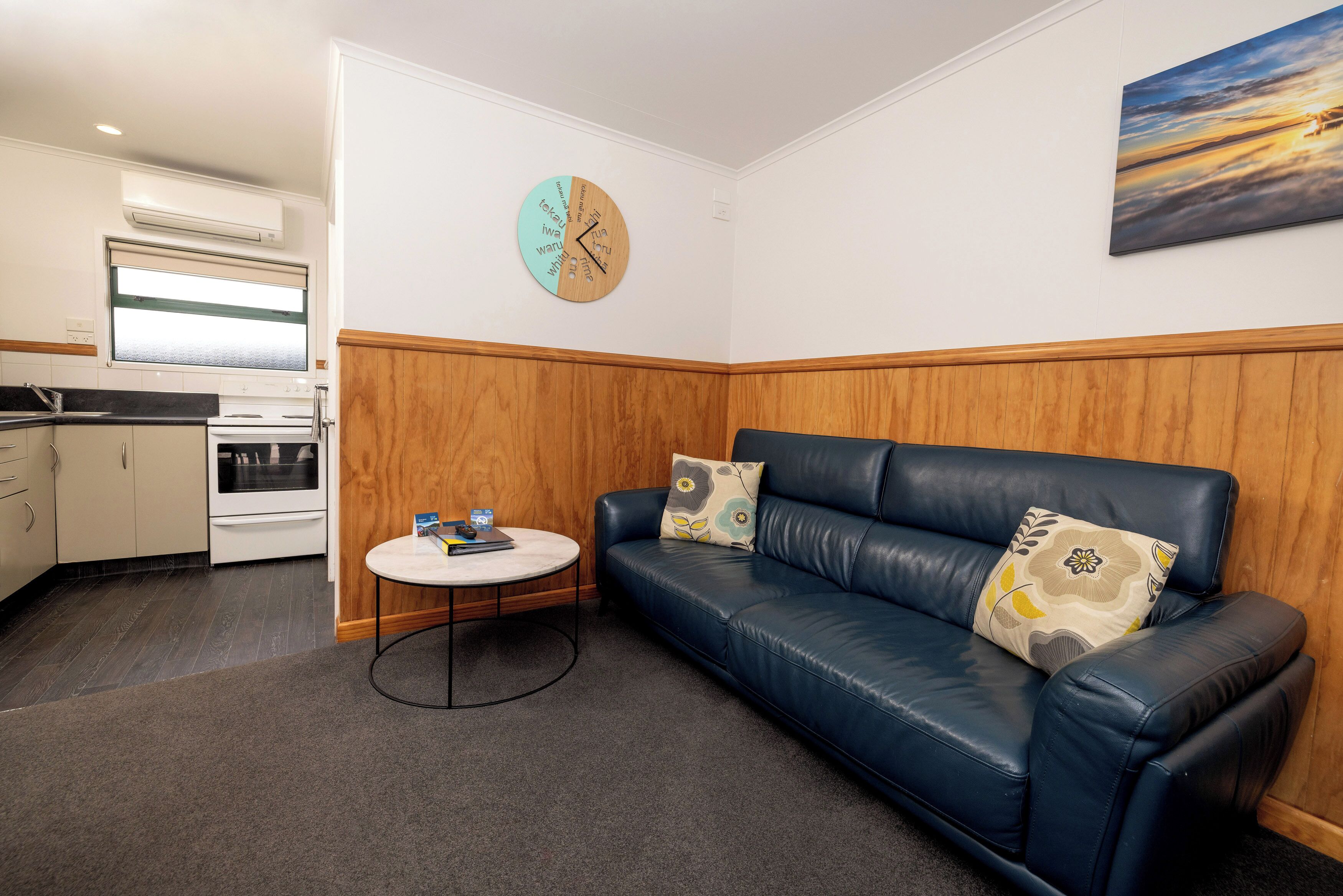 A compact living space with a kitchenette, a blue leather sofa with floral cushions, a round coffee table, and a clock on the wall.