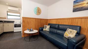 A compact living space with a kitchenette, a blue leather sofa with floral cushions, a round coffee table, and a clock on the wall.