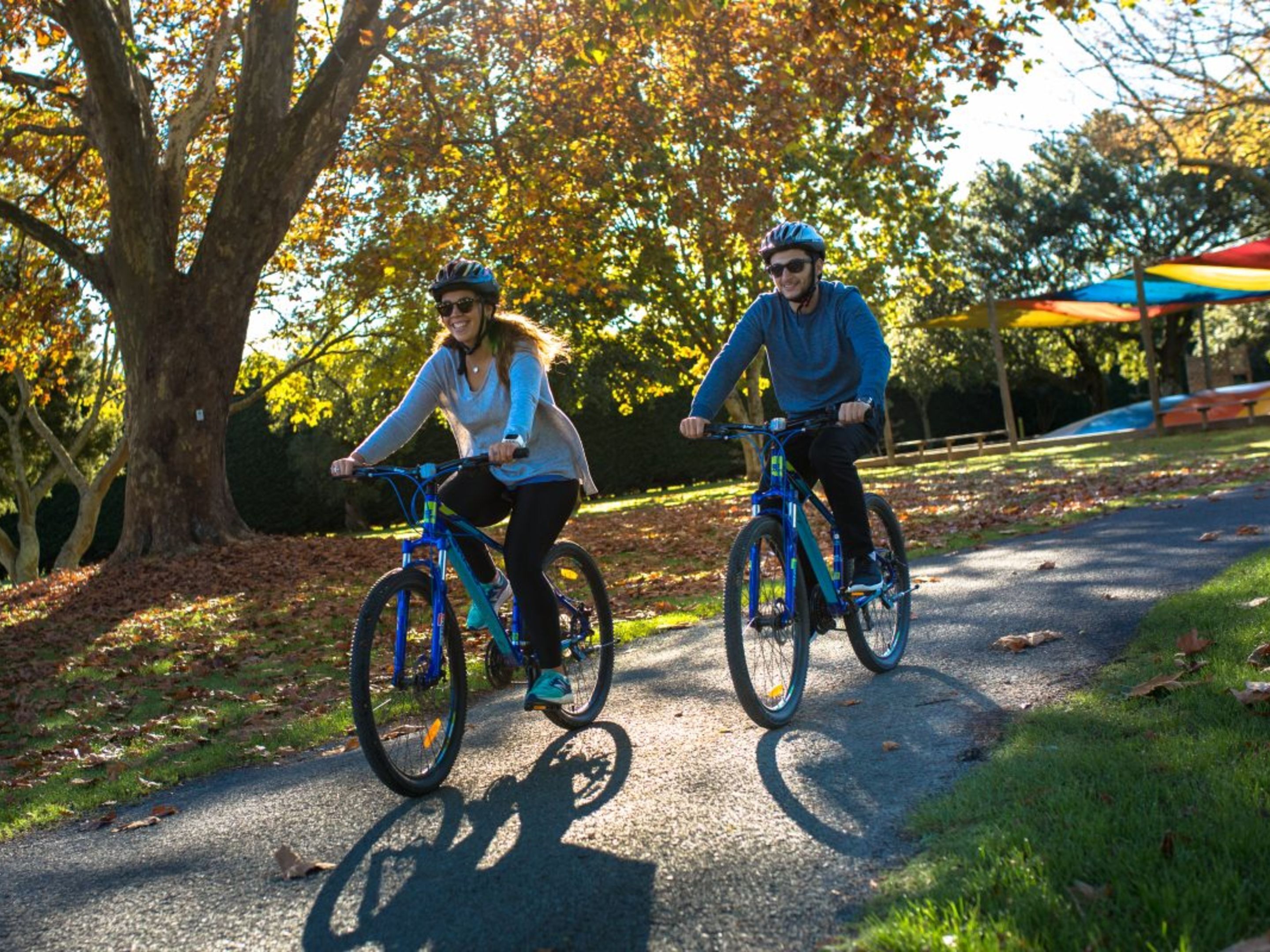 Twee personen fietsen op een verhard pad in een park, met bomen en een kleurrijke paraplu op de achtergrond.