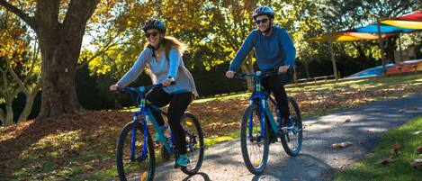 Twee personen fietsen op een verhard pad in een park, met bomen en een kleurrijke paraplu op de achtergrond.