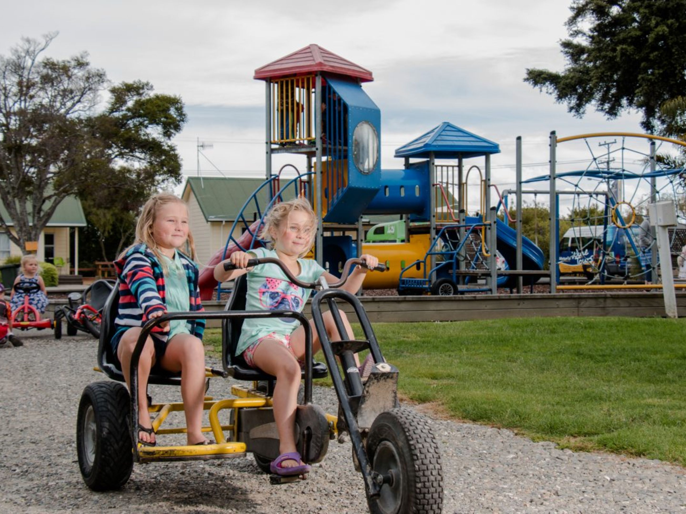 Two children on a ride-on vehicle at a playground with various play structures in the background.