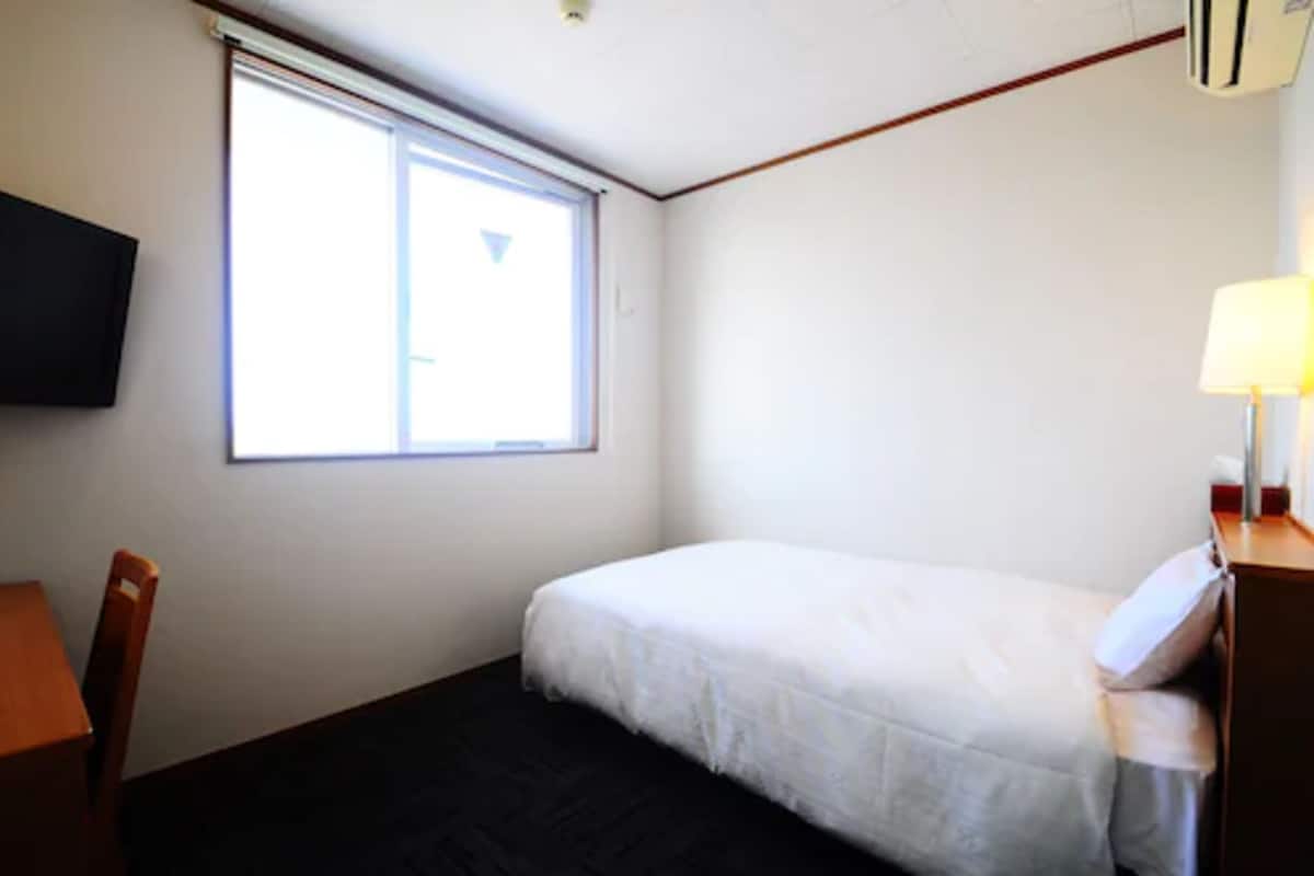 Een kleine, schone kamer met een enkel bed, een bureau, een stoel, een aan de muur bevestigde televisie en een raam voor natuurlijk licht.