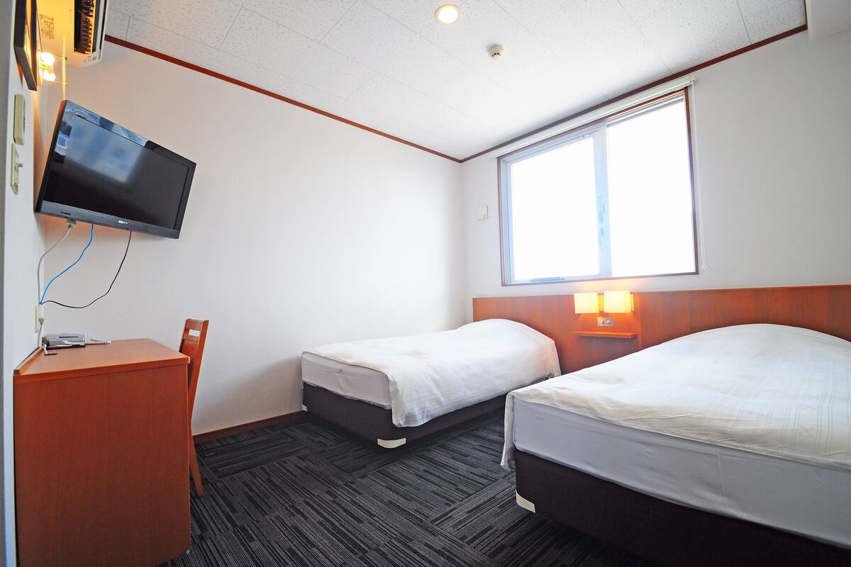 Een hotelkamer met twee enkele bedden, een bureau met stoel, een flatscreen televisie aan de muur en een raam dat voor natuurlijk licht zorgt.