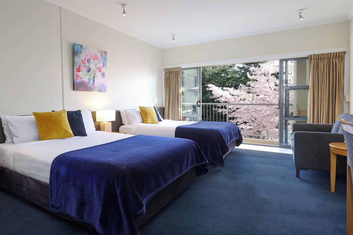 A hotel room with two beds, a balcony with a view of cherry blossoms, and a chair.