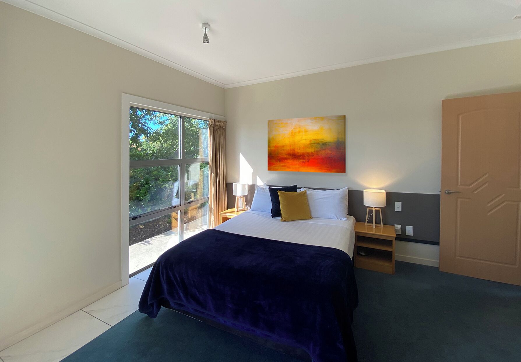 A modern hotel room with a large bed, two bedside tables, a window with curtains, a door, and a painting on the wall.