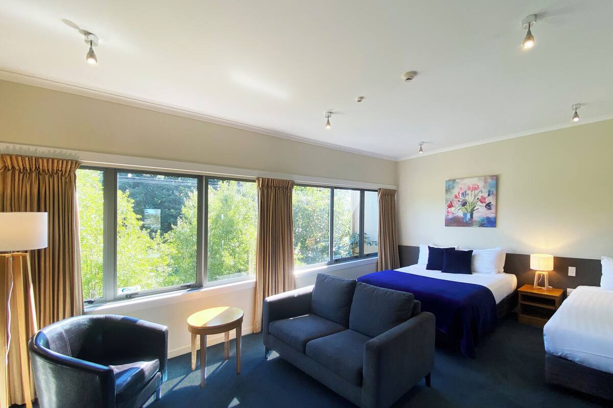 A hotel room with a bed, a sofa, a small table, and a chair. There are windows with curtains, a painting on the wall, and ceiling lights.