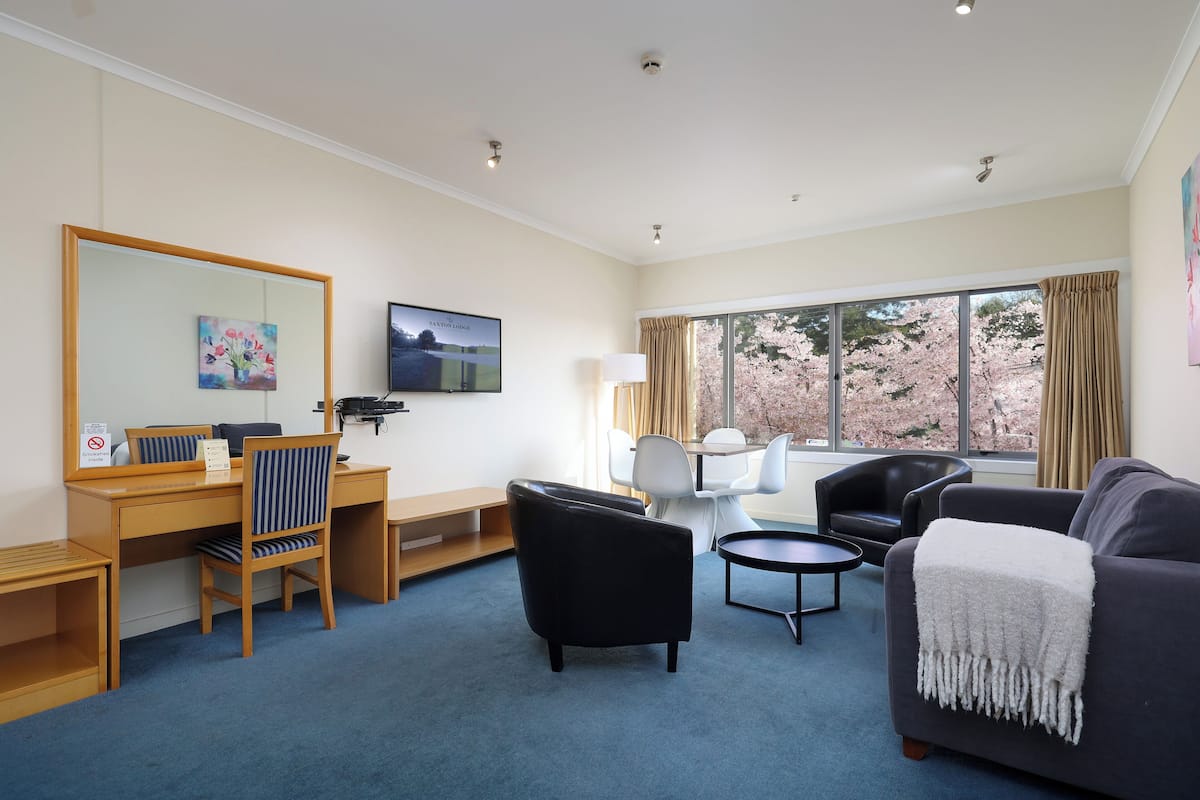 A hotel room with a desk, a black armchair, a grey sofa, a small table, and a large window with curtains.