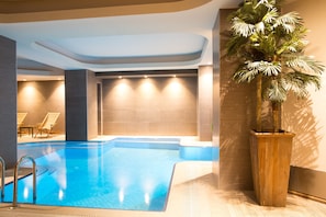 An indoor swimming pool with clear blue water, surrounded by tiled walls and a wooden planter with a palm tree. - Art Deco Suites Istanbul (Istanbul)