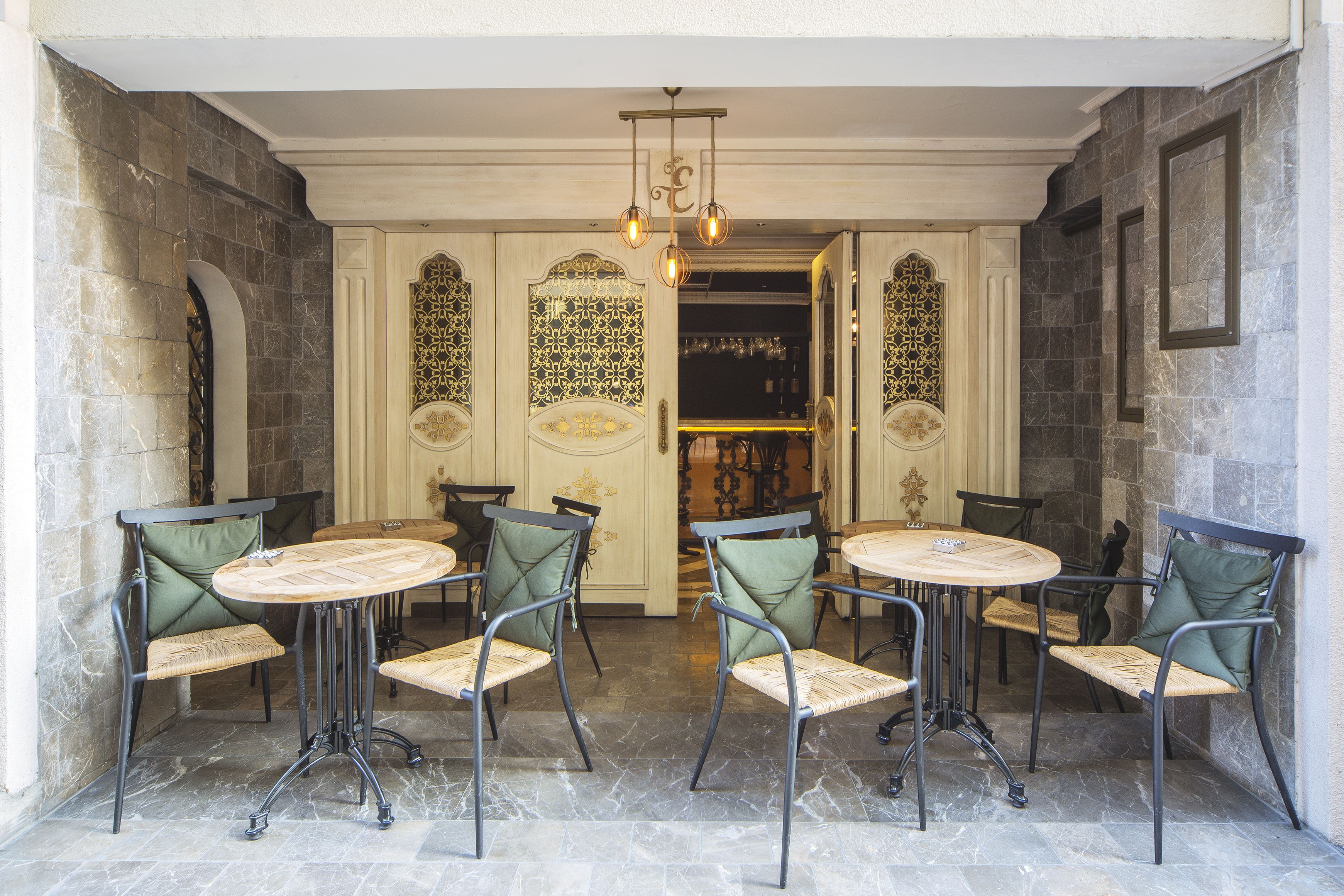 A dining area with four round tables and chairs, each with a woven seat cushion and metal frame. The walls are adorned with intricate patterned designs, and a chandelier hangs from the ceiling.