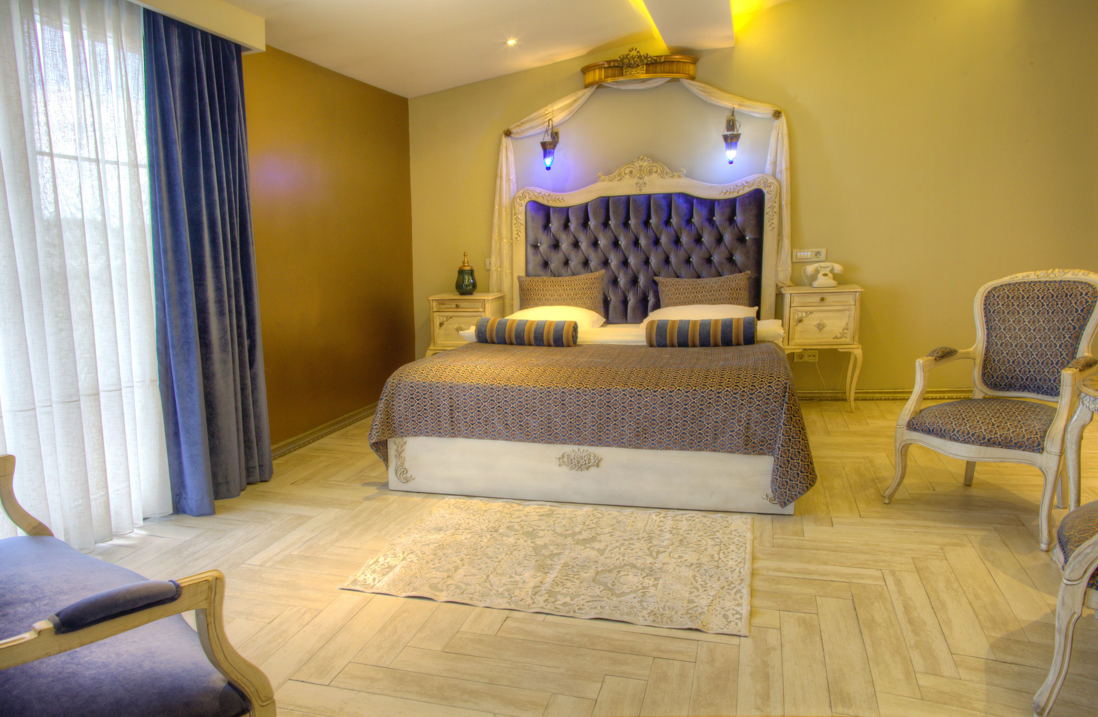 A large bed with a tufted headboard, two bedside tables, a chair, and a large window with curtains.