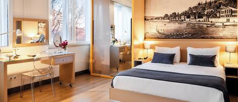 Una habitación de hotel moderna con una cama grande, un escritorio con una silla, un espejo y un cuadro de una escena junto al río.