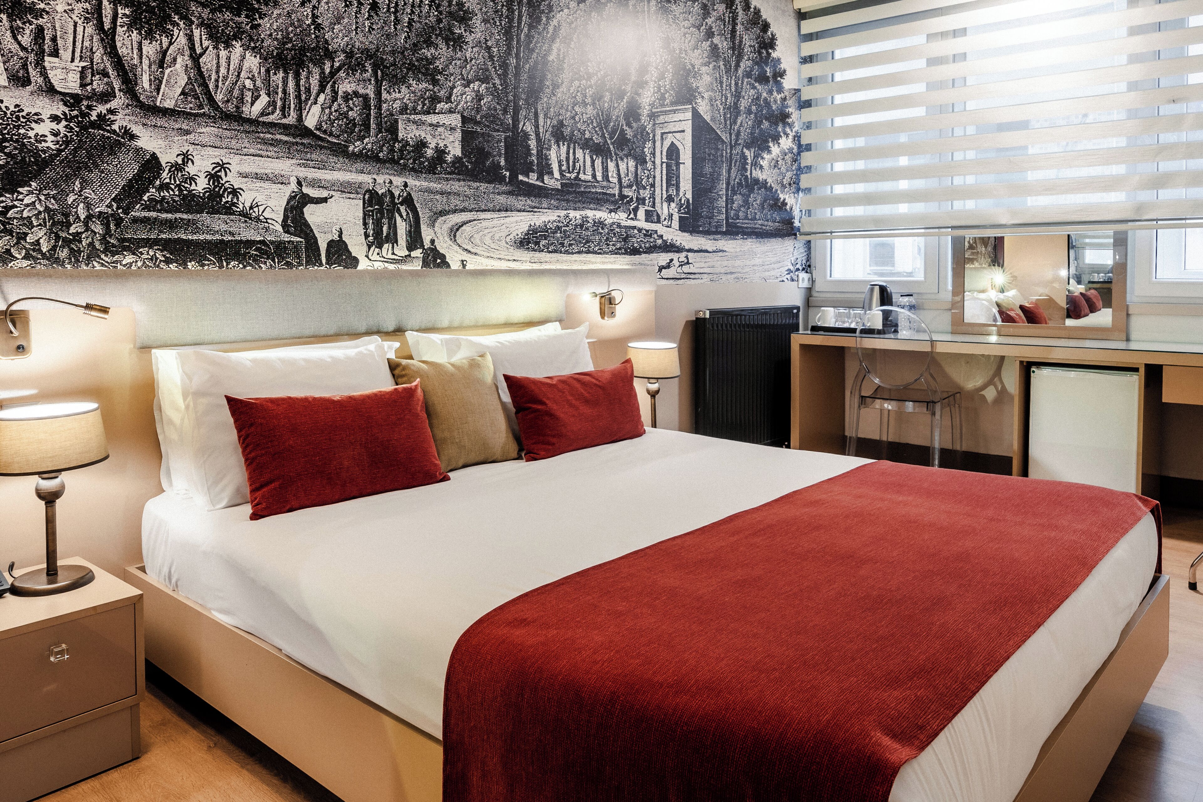 A neatly made bed with a red blanket, two bedside lamps, and a large wall mural depicting a historical scene.
