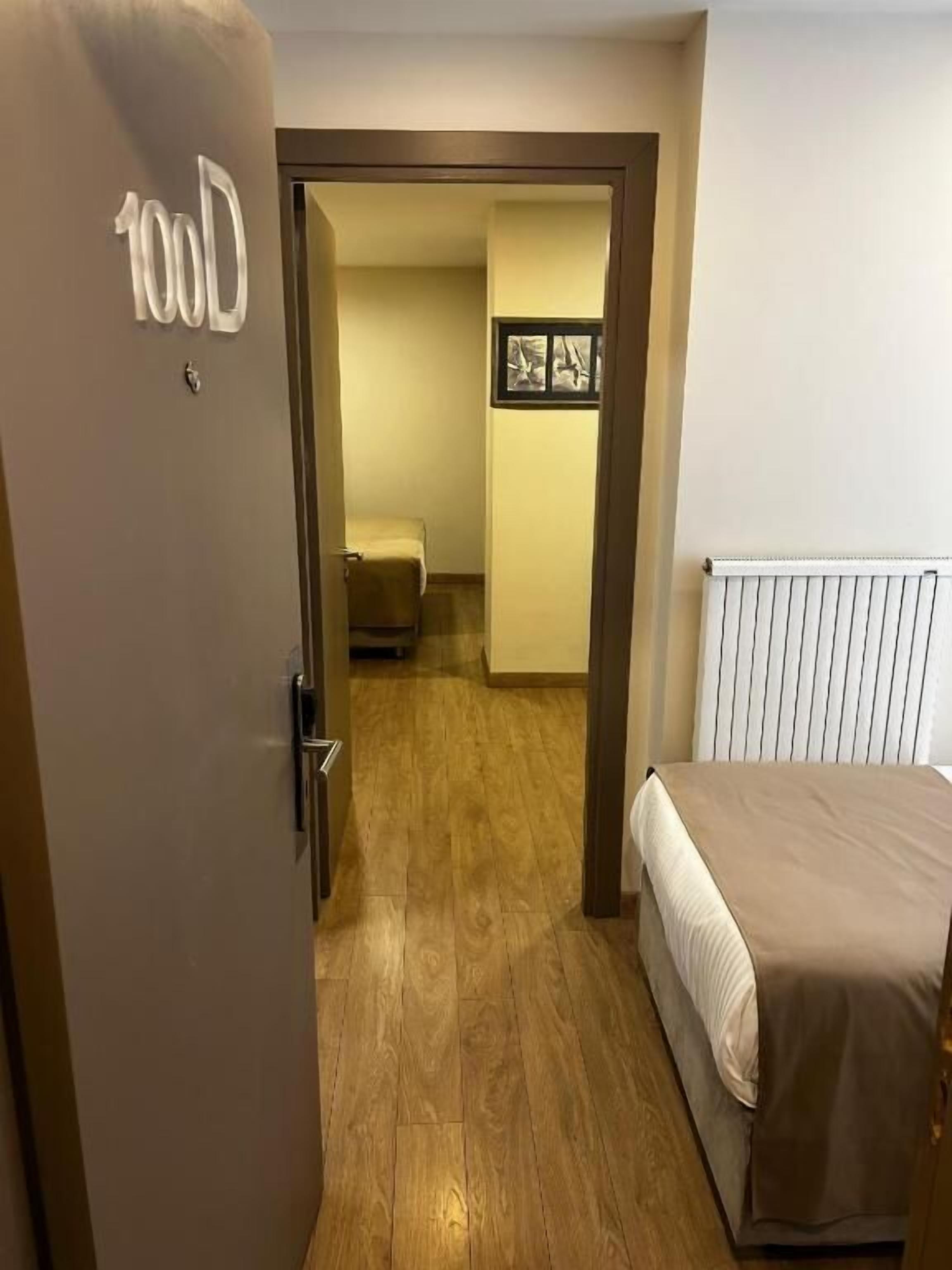 Habitación de hotel con una cama, una puerta con un número, un radiador y un cuadro en la pared.
