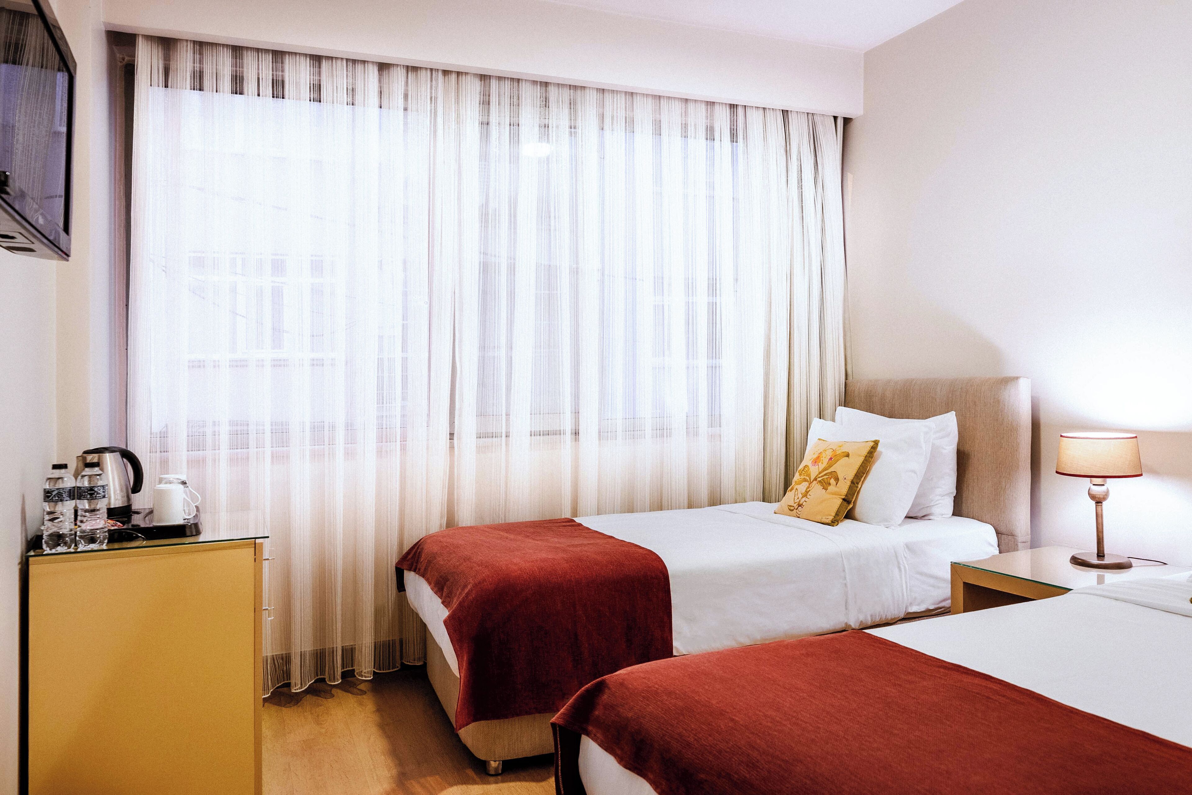 A hotel room with two beds, a desk, a lamp, and a window with sheer curtains.
