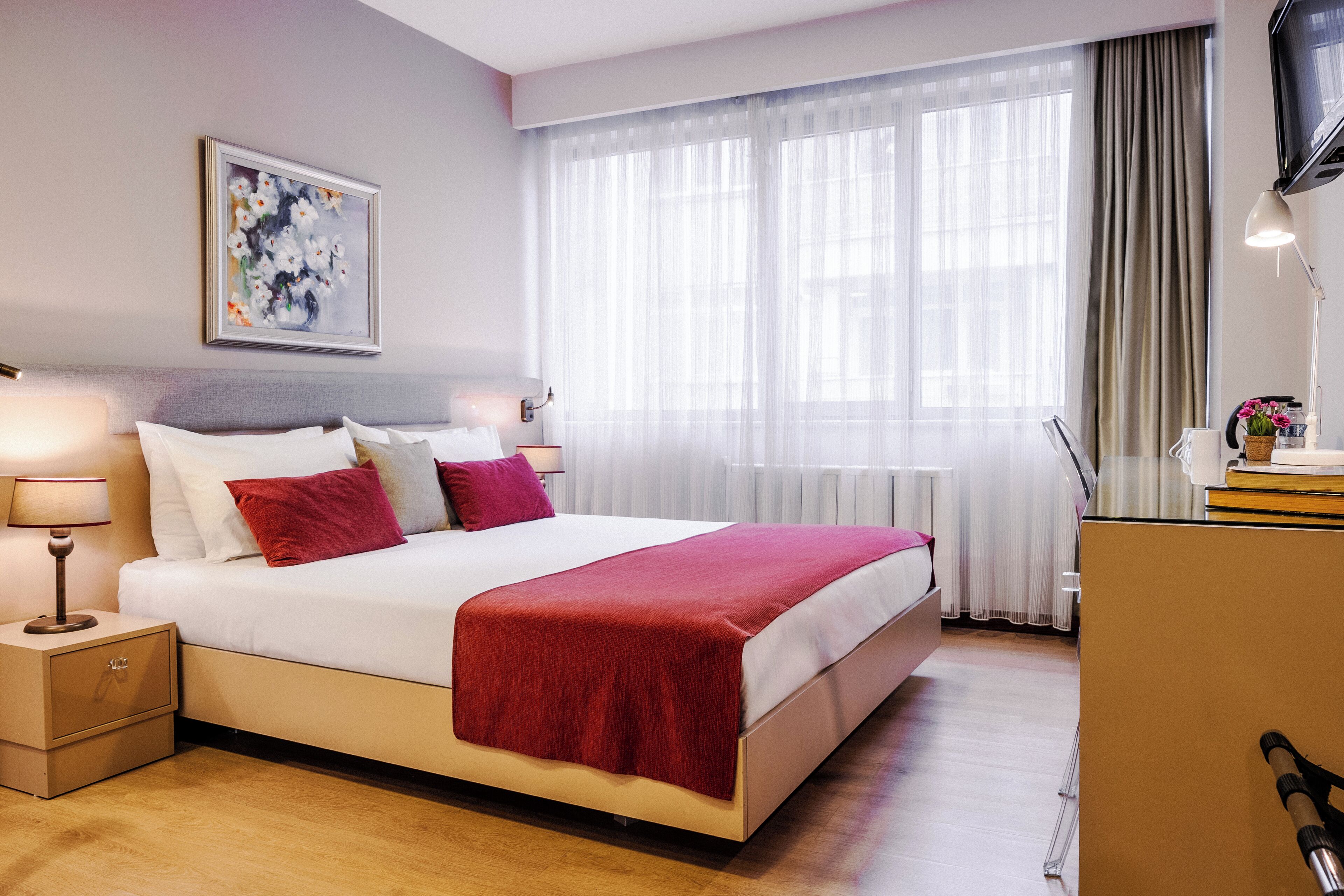 Une chambre d’hôtel bien aménagée, avec un grand lit, une table de chevet avec une lampe, une œuvre d’art encadrée au mur et une fenêtre ornée de rideaux légers.