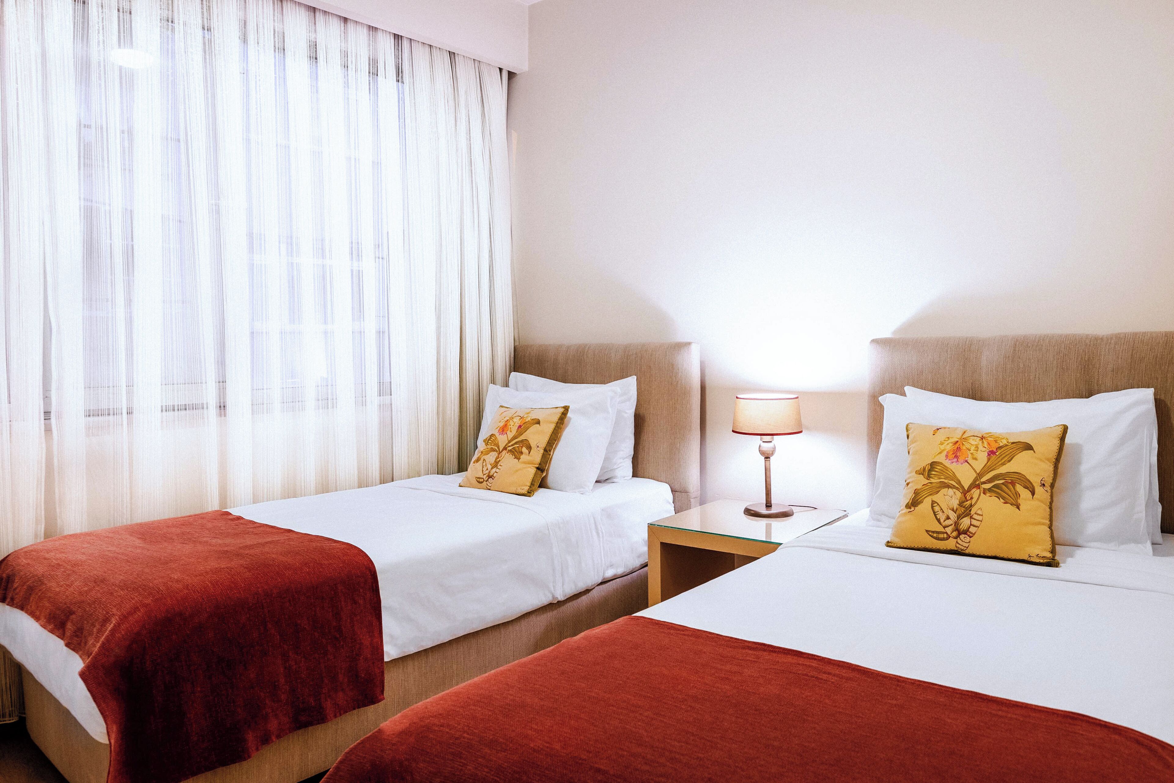 A hotel room with two beds, each with a red blanket and a floral pillow. There is a nightstand with a lamp between the beds.