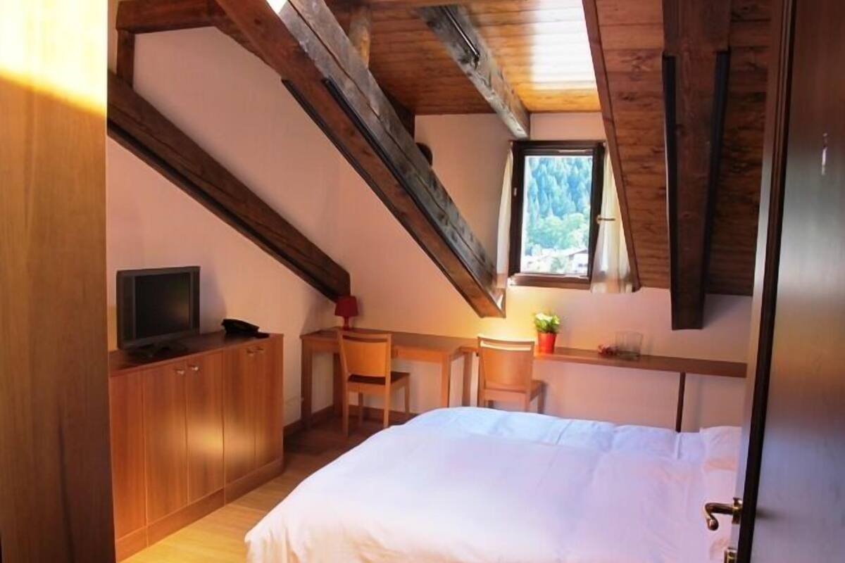 A bedroom with a bed, a desk, a chair, a television, and a window with a view of trees.