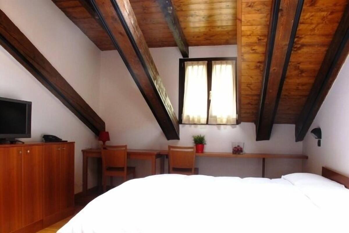 A bedroom with a wooden ceiling, a bed, a desk, a chair, a television, and a window with curtains.