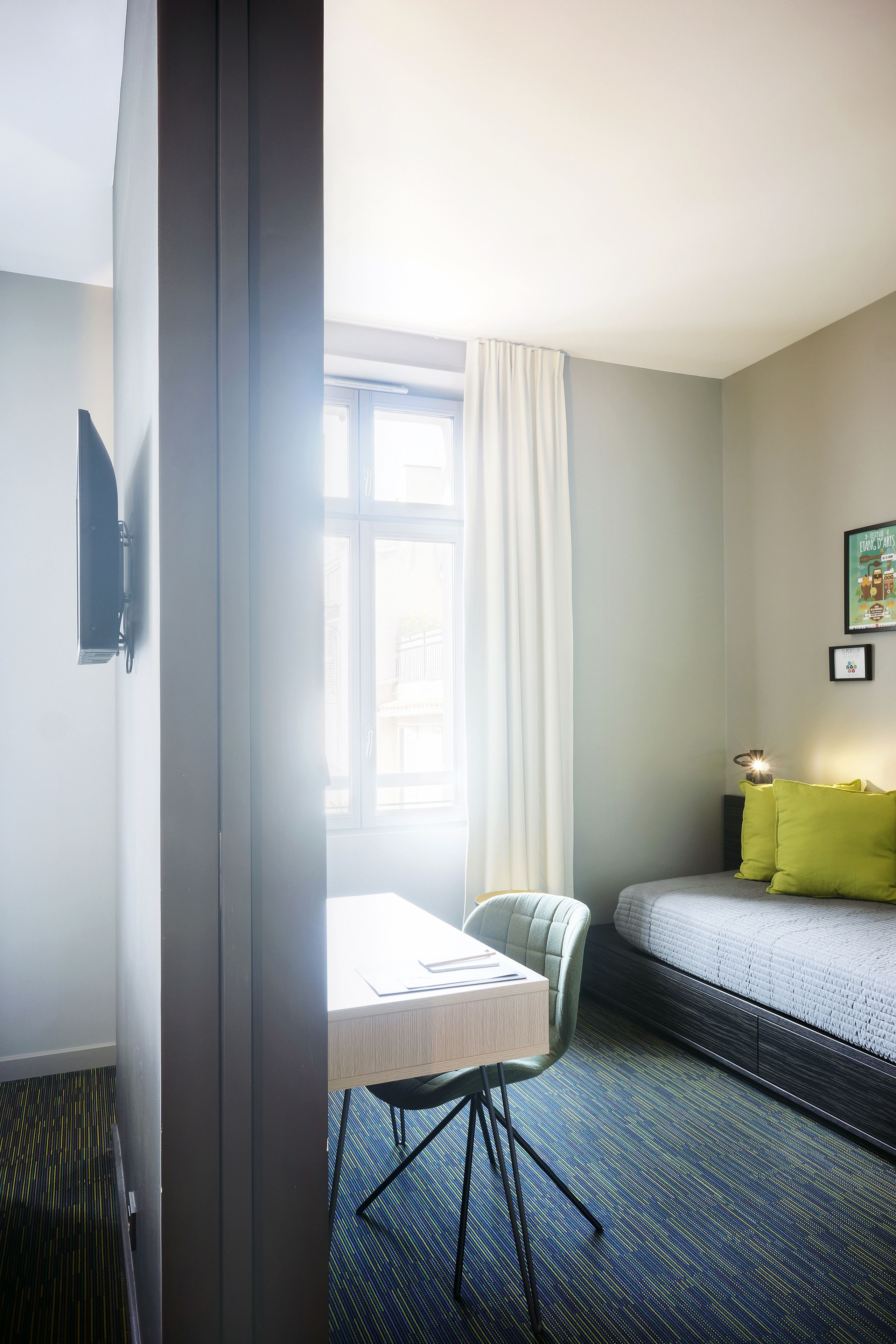 Quarto de hotel com cama, escrivaninha com cadeira, janela com cortinas e televisão na parede.