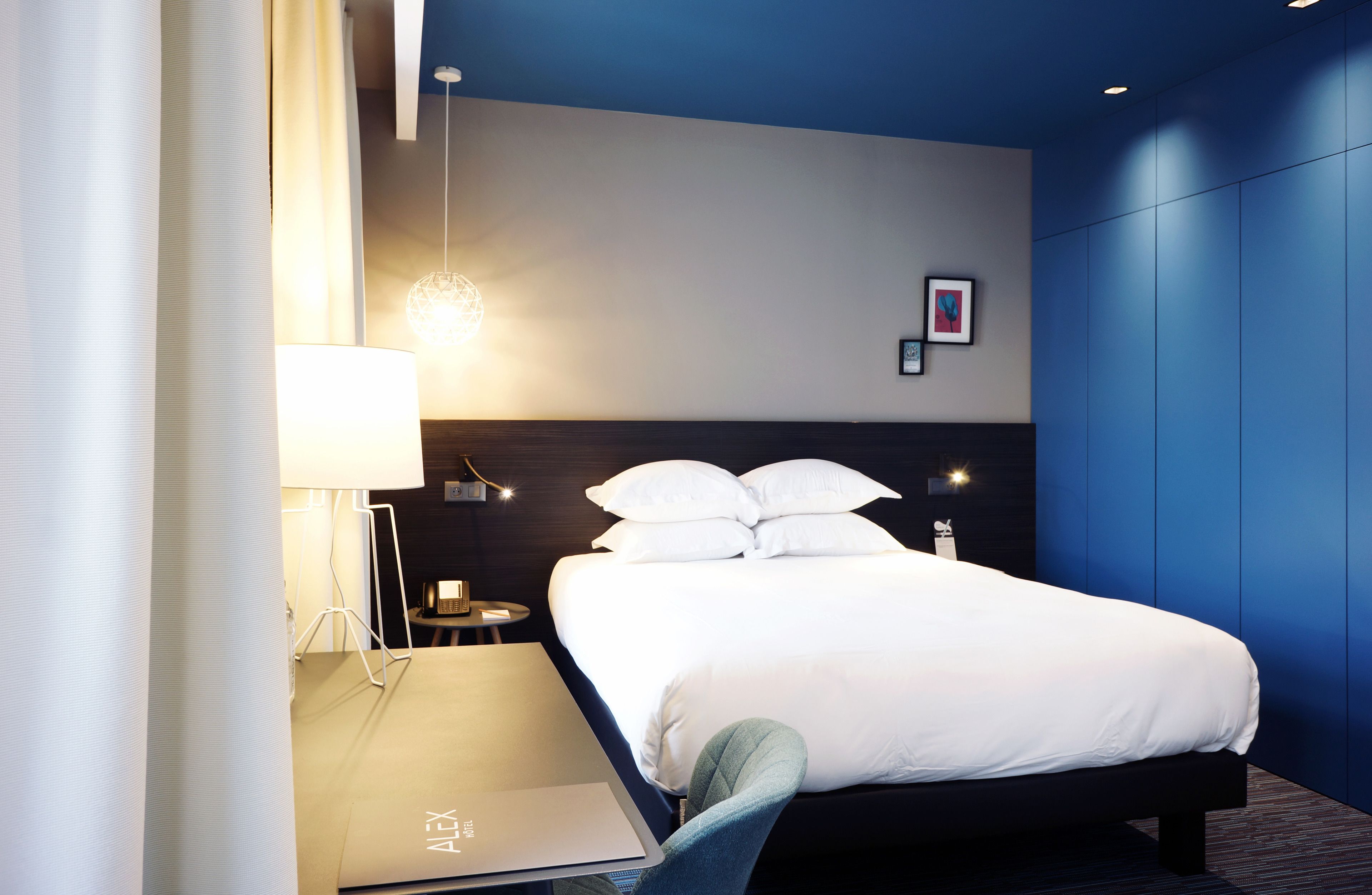 A modern hotel room with a large bed, a desk with a laptop, a lamp, and a picture on the wall.