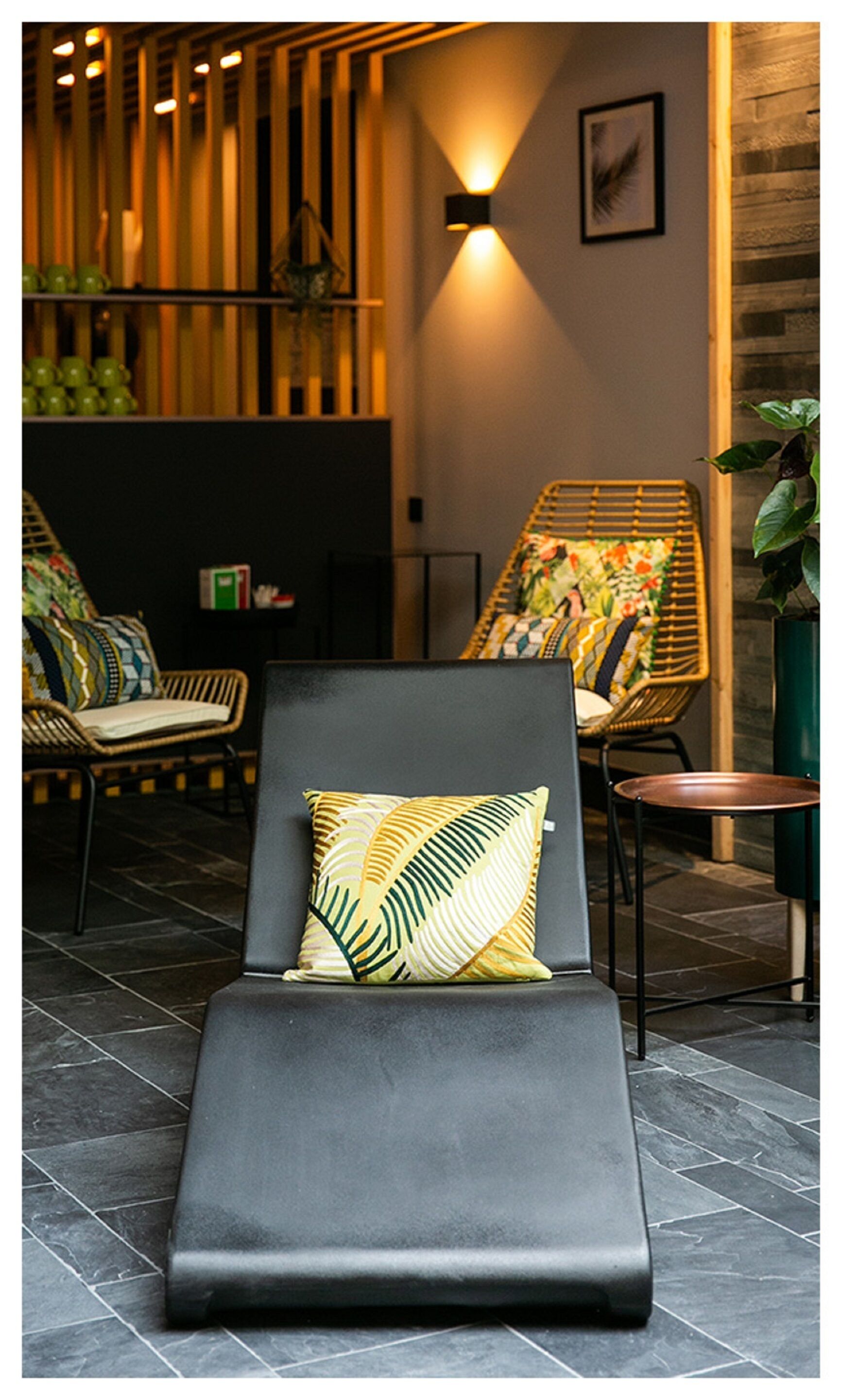A modern lounge area with a black lounge chair, a wicker chair with a colorful cushion, and a small table with a green vase.