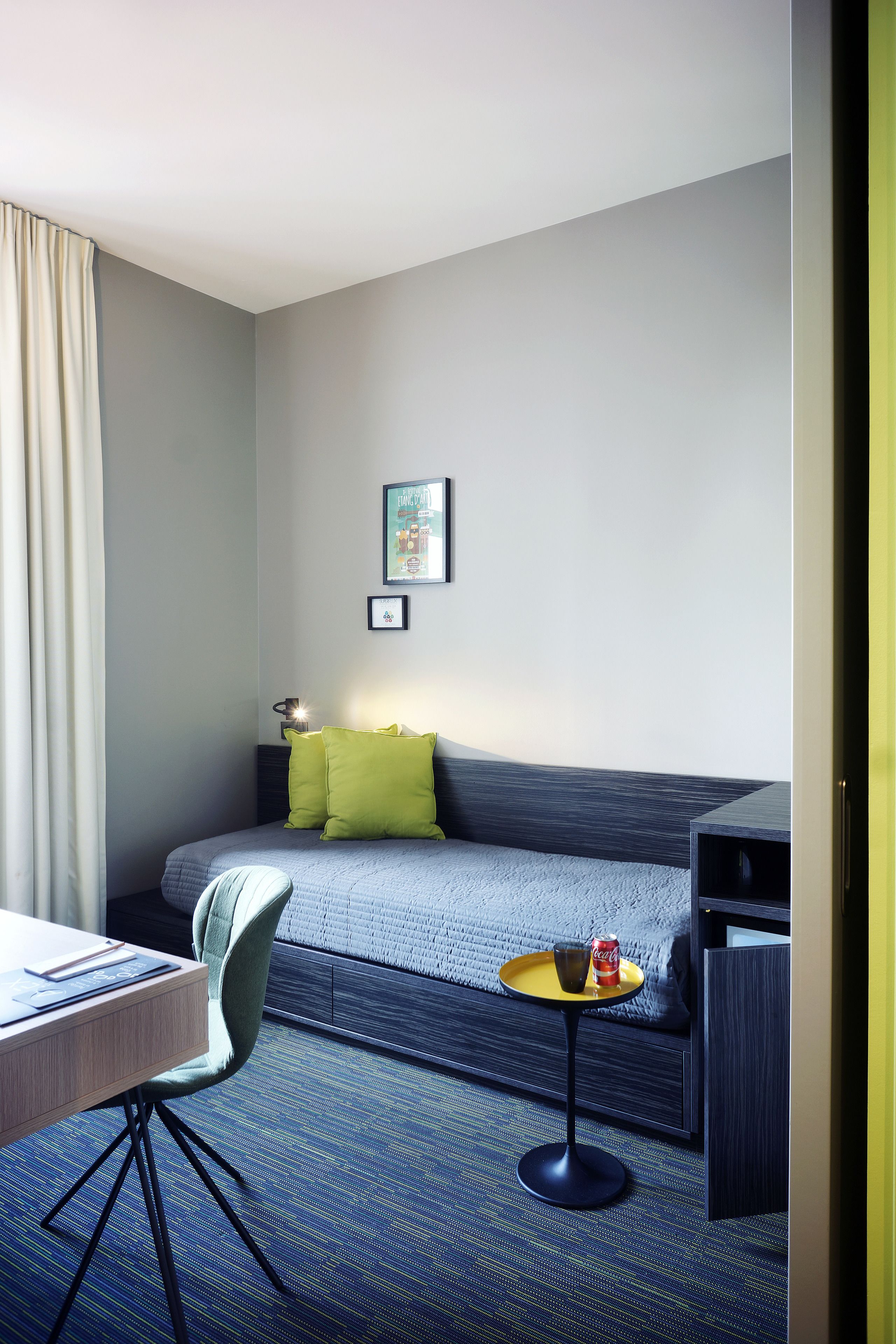 A modern hotel room with a bed, a desk, a chair, a small table with a drink, and a sofa.