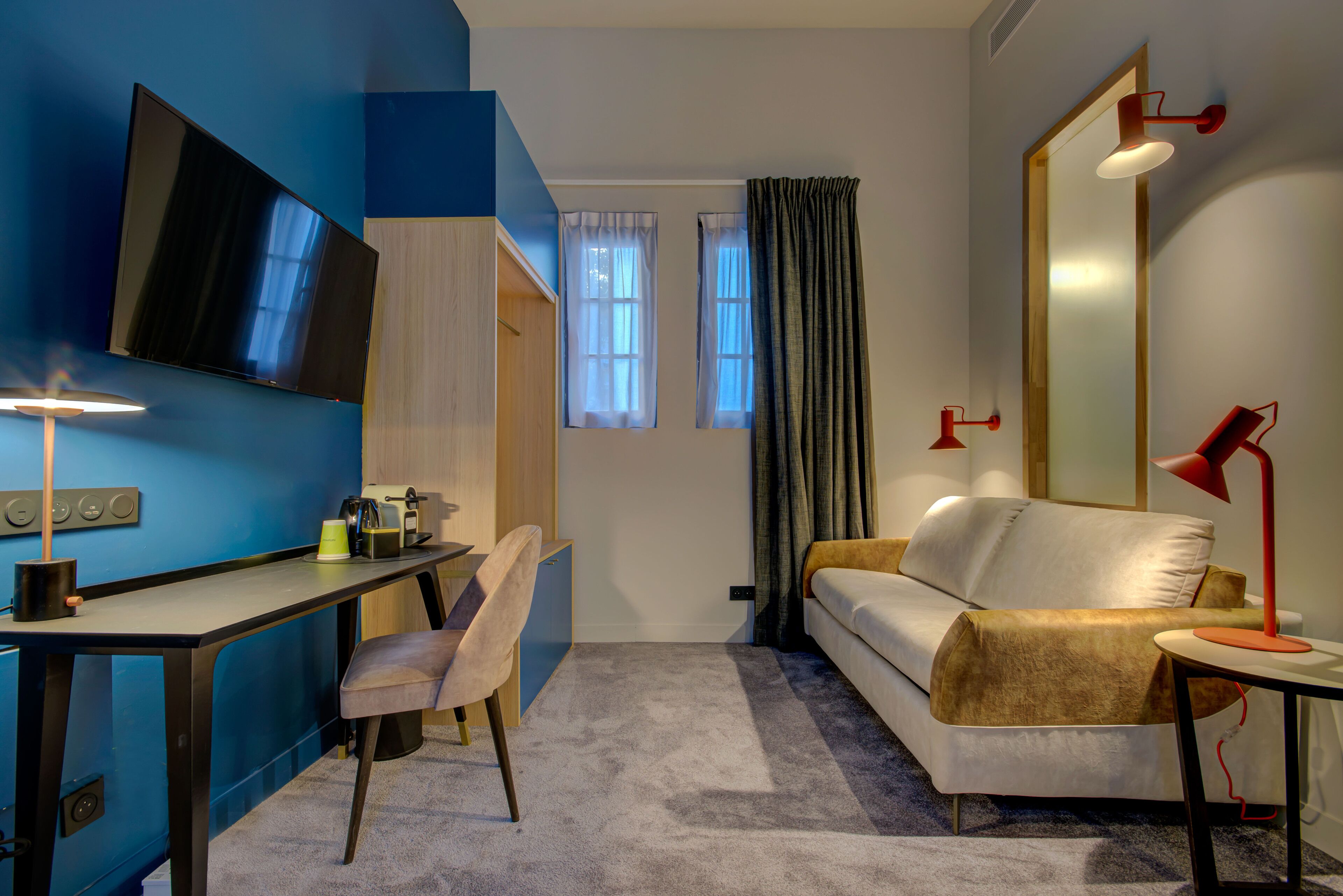 A modern hotel room with a desk, chair, sofa, and a large mirror.