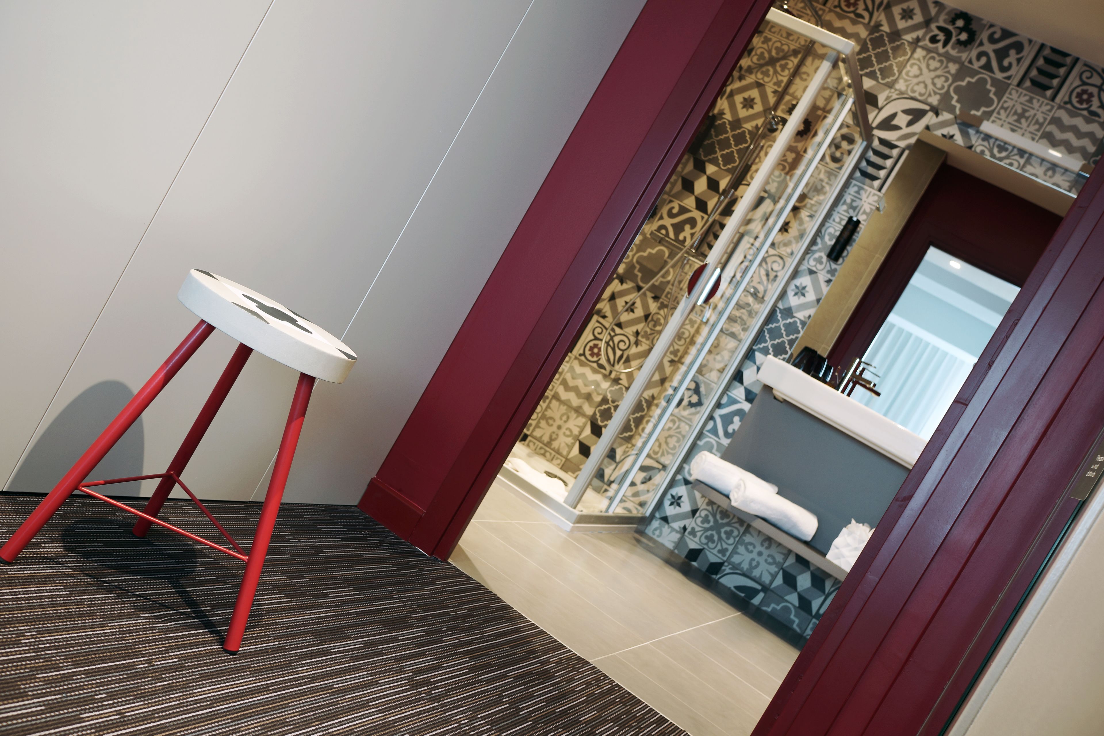 Un baño moderno con baldosas estampadas, un lavamanos blanco y un taburete rojo.