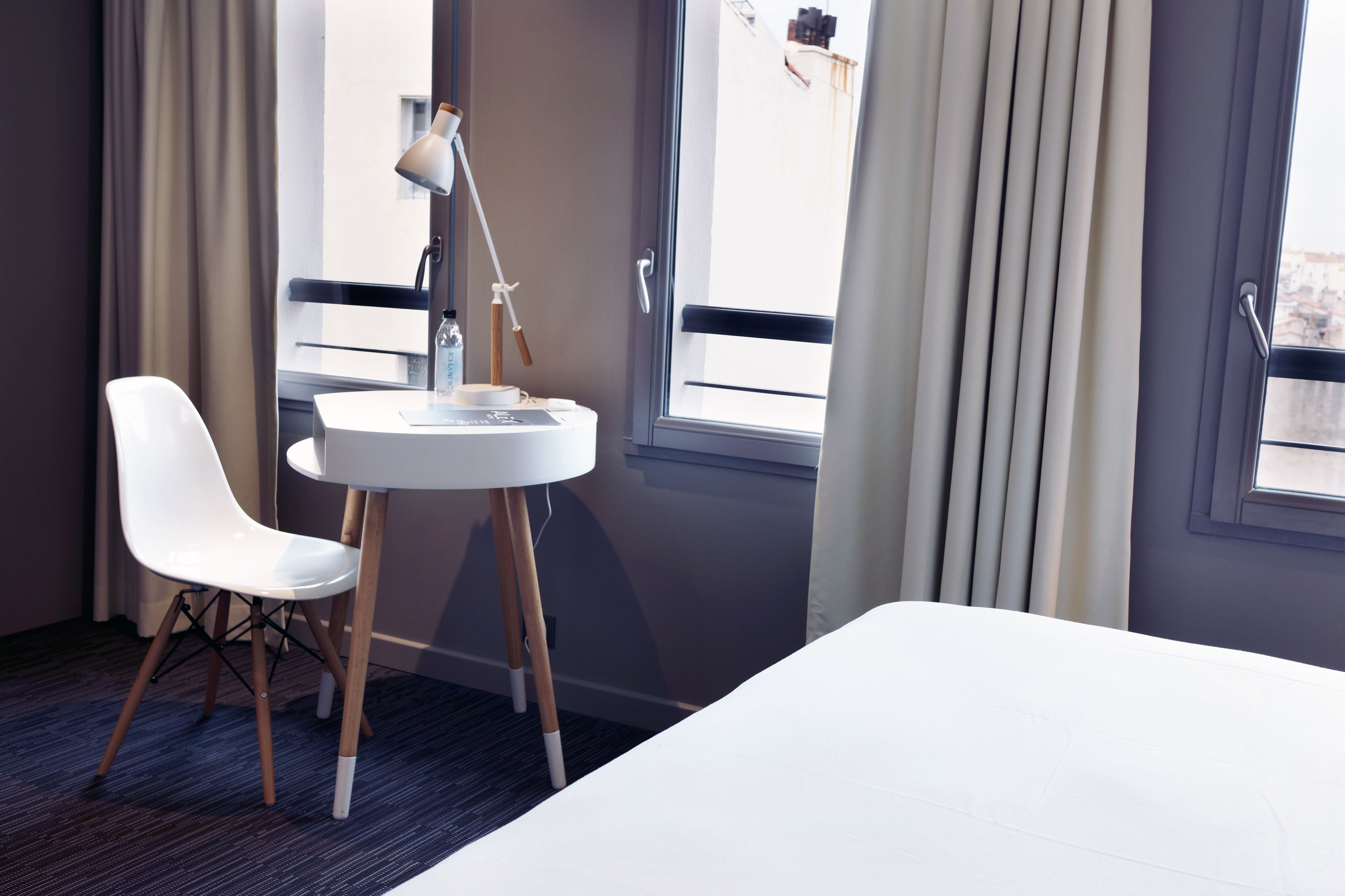 Une chambre d’hôtel avec un lit, une chaise blanche, une table ronde avec une lampe et une fenêtre avec des rideaux.
