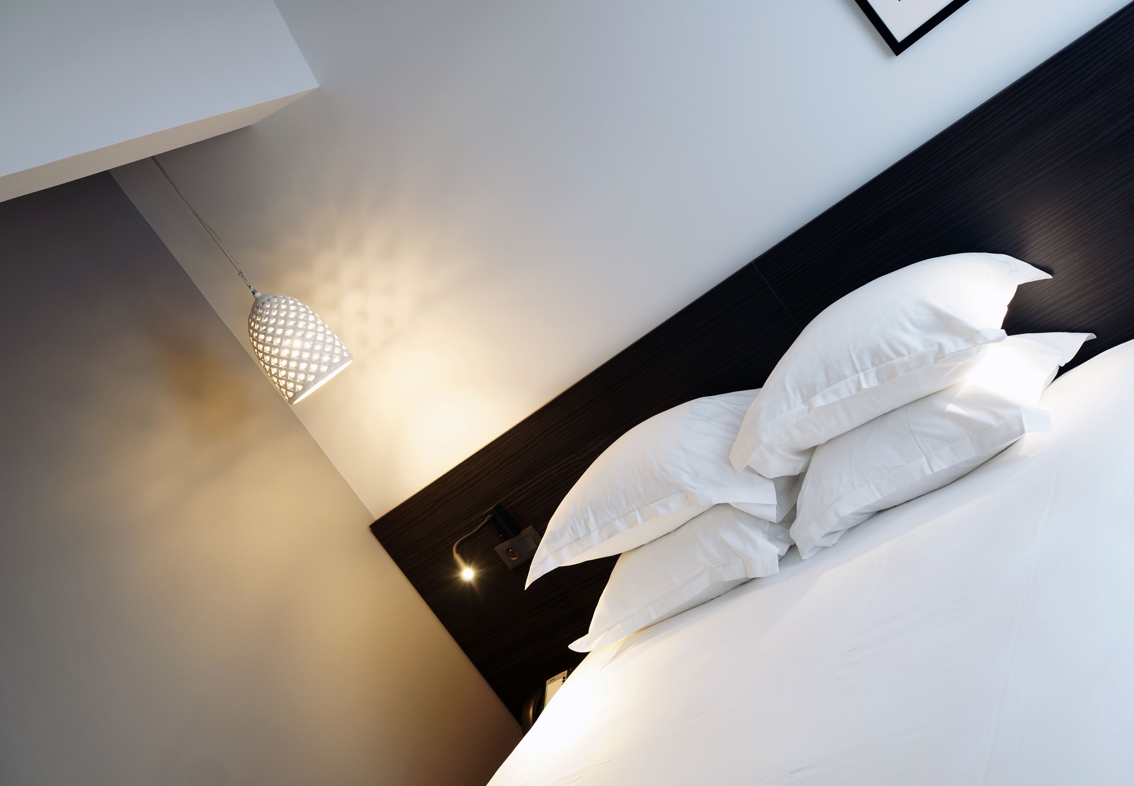 Un lit bien fait, avec du linge de lit blanc et des oreillers, une suspension lumineuse et un tableau encadré au mur.